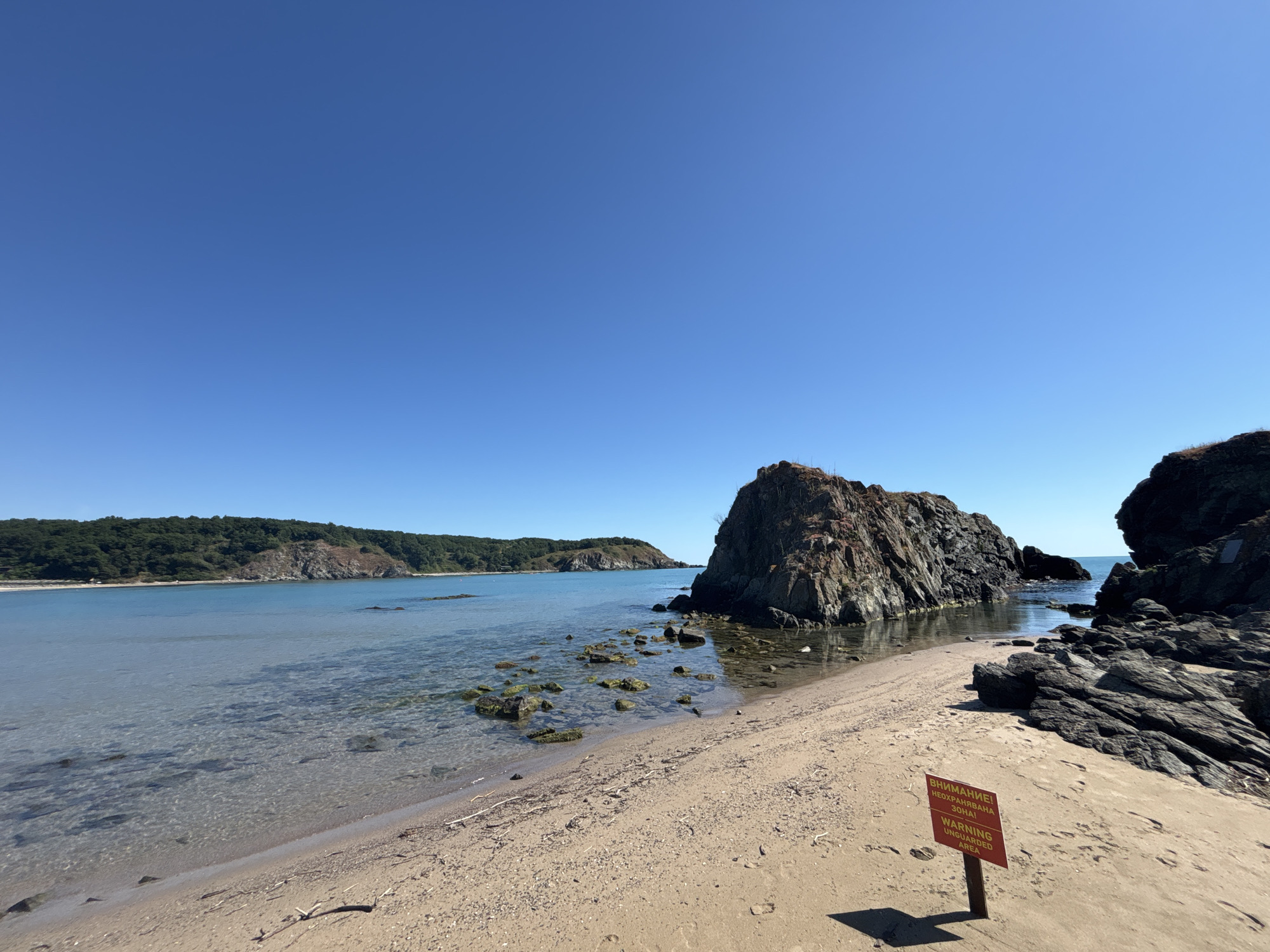 This is a scenic photograph of a beach on a clear, sunny day. The foreground features a sandy beach with some driftwood scattered on it. A small red sign with yellow lettering is planted in the sand, displaying the text "ВНИМАНИЕ! НЕОХРАНЯВАНА ЗОНА! WARNING! UNGUARDED AREA". To the right of the sign, dark, rugged rocks rise from the water and shoreline. In the middle ground, clear, calm water laps at the beach, with submerged rocks visible beneath the surface and a larger rock formation jutting out of the sea. In the background, a lush, green hillside slopes down to the water, suggesting a coastal landscape. The sky is a vast, unbroken expanse of clear blue. This scene appears to be located in Rezovo, Bulgaria, a coastal town known for its natural beauty.
