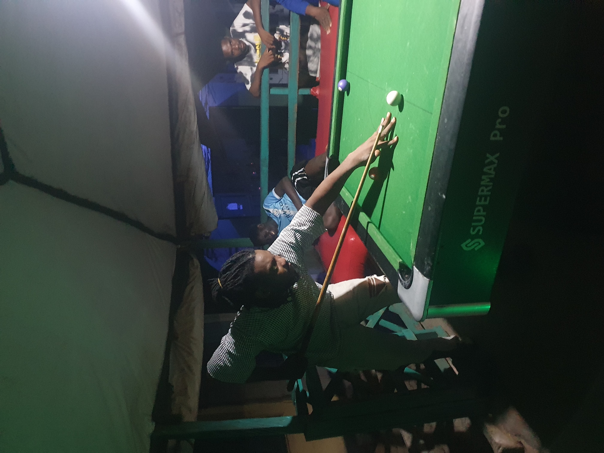 The image depicts a nighttime scene at an outdoor pool hall, possibly located in Lokoja, Nigeria, under a covered structure. A Black male, wearing a checkered shirt and light-colored pants, is actively playing pool, positioned over the green felt table. He is in the process of striking a white cue ball with a pool cue. Other individuals are visible in the background, some appearing to be watching the game. The environment is dimly lit, with the primary light source seemingly coming from above the pool table and the general ambiance suggesting a casual, social gathering. The pool table itself is branded with "SUPERMAX Pro."
