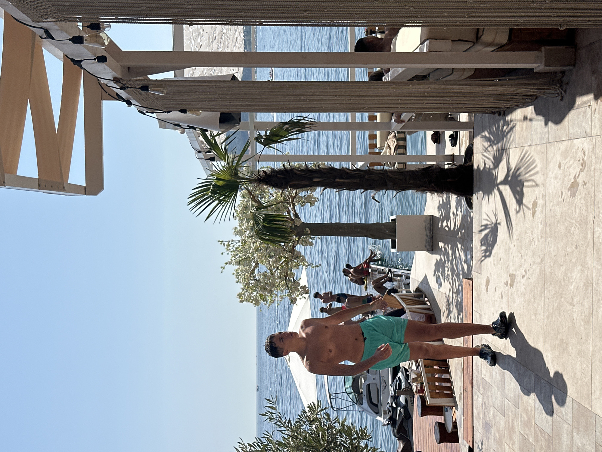 The image depicts a sunny day at a waterfront establishment in Budva, Montenegro. In the foreground, a young man wearing turquoise swim trunks and black slip-on shoes walks on a tiled patio. Behind him, people are visible on a wooden dock extending into the blue sea. Several individuals are seated on the dock, some appearing to be relaxing, while others are standing. A white umbrella provides shade over a section of the dock. A small white boat with jet skis parked nearby is also present.
To the right of the foreground subject, a covered outdoor seating area is visible with white cushioned furniture and decorative rope curtains. String lights hang from the ceiling structure of this area. A tall palm tree with a thick trunk and fronds stands between the tiled patio and the water. Further in the background, the clear blue sky meets the calm sea. In the distance, a few boats are visible on the water. The overall scene suggests a leisure atmosphere at a beachside resort or restaurant.