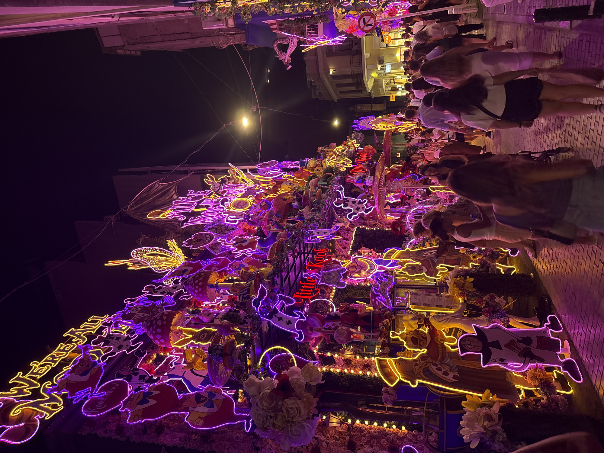 The image depicts a bustling street scene at night in Athens, Greece, specifically on Adrianou Street in the Plaka district. The primary subject is a building facade heavily decorated with vibrant neon lights, predominantly in purple and yellow, forming characters and motifs from "Alice in Wonderland." Notable illuminated elements include the phrase "WE'RE ALL MAD HERE," various playing cards, mushrooms, the Cheshire Cat, and other figures from the story. The decorations extend along the building's edge and spill onto the sidewalk.
A crowd of people, casually dressed, are walking along the cobblestone street, some looking at the decorations, others engaged in conversation. The lighting from the neon signs casts a strong purple and pink glow on the street and the people. Several shops and buildings line the street, with some appearing to be restaurants or bars, indicated by outdoor seating and lighting. A street sign with a red circle and line, possibly indicating a no-entry or no-parking zone, is visible on the right side. The overall atmosphere is festive and lively, characteristic of a popular tourist area at night.