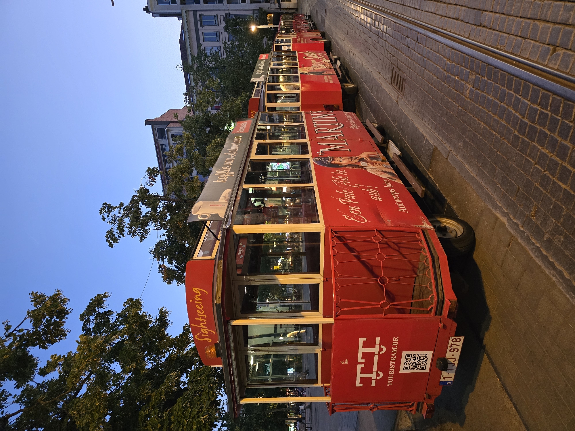 A red, open-air tourist tram sits on tracks in Antwerp, Belgium. The tram is decorated with advertisements for "Sightseeing," "Martini's," and "Koffie van Antwerpen." A QR code and license plate "1-IJJ-978" are visible on the back of the tram. The tram is lined up with other similar trams. The surrounding environment includes cobblestone streets, trees, and buildings. The sky is a deep blue, indicating it is either early morning or late evening.
