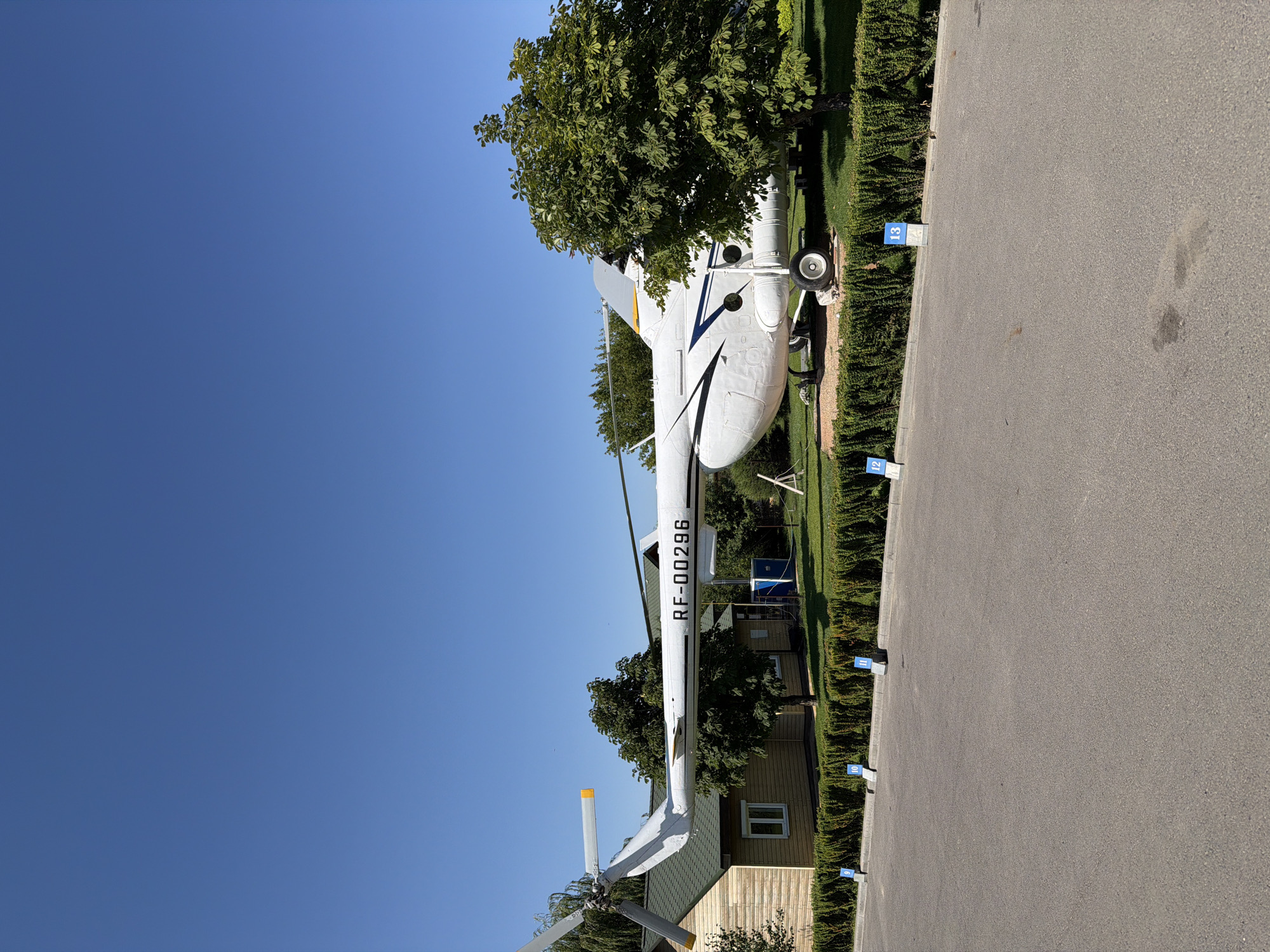 This is a full, eye-level, outdoor, natural light photograph of a white helicopter on display. The helicopter is positioned horizontally and slightly angled towards the right side of the frame. It has the registration markings "RF-00296" in black text on its side. The helicopter is in front of a long, low-rise building with a green roof and wooden siding. A row of manicured green bushes borders the area in front of the building and the helicopter. Beyond the bushes and helicopter, there are several trees. In the foreground, there is an asphalt surface with parking markers numbered 9, 10, 11, 12, and 13 visible along the edge of the bushes. The sky is clear and blue. The scene is set in the city of Kamyshin, Russia, likely at an aviation museum or park, given the static display of the helicopter and the context of its historical significance.