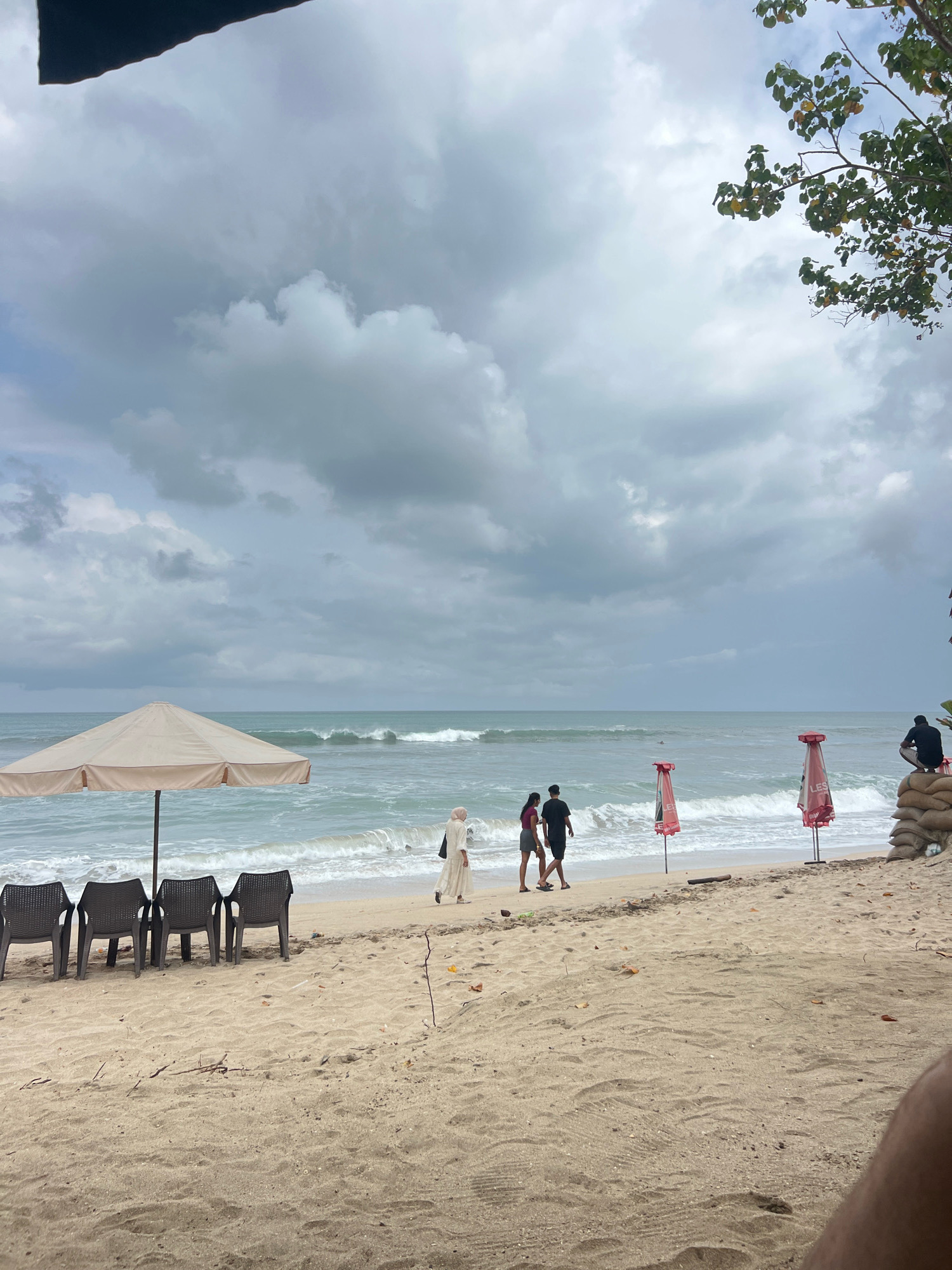 The image depicts a beach in Kuta, Indonesia, under a cloudy sky. In the foreground, the sandy beach is sparsely populated, with four empty plastic chairs arranged under a large, cream-colored umbrella. Further back, a couple walks along the water's edge. To their right are two red beach umbrellas, and to the far right, a person sits atop a stack of sandbags, overlooking the ocean. The sea is active with moderate waves. The overall atmosphere is tranquil, despite the overcast weather.