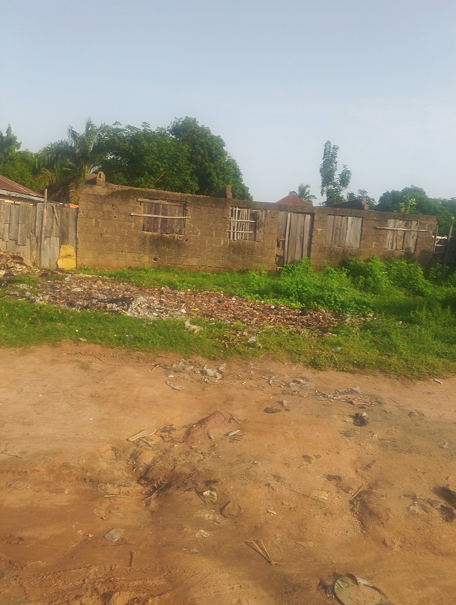 The image shows an outdoor scene in Jalingo, Nigeria. In the foreground, there is a dirt road with scattered debris. Behind the road, a patch of overgrown grass is visible, also littered with trash. Further back, an unfinished or derelict building made of cinder blocks stands. This structure has wooden planks covering its windows and doors, suggesting disuse. A wooden fence runs along the left side of the building. The background features lush green trees and a clear sky. The overall impression is one of an unmaintained urban or semi-urban environment.