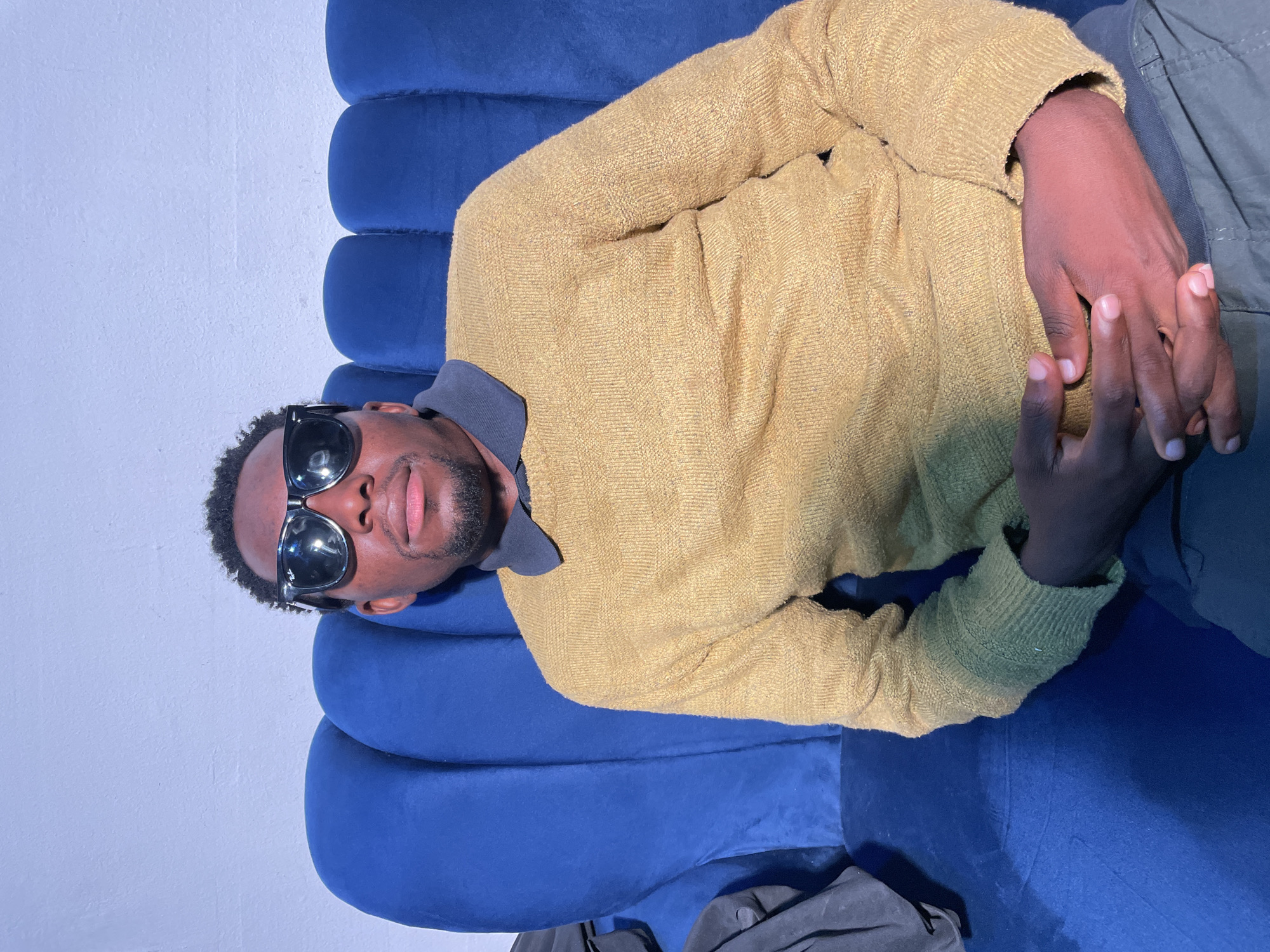 A dark-skinned man lies on a blue couch. He is wearing a yellow sweater and sunglasses. He is in an indoor setting. Location: Barakin Akawo, Nigeria is not explicitly provided and cannot be determined from visual information alone.