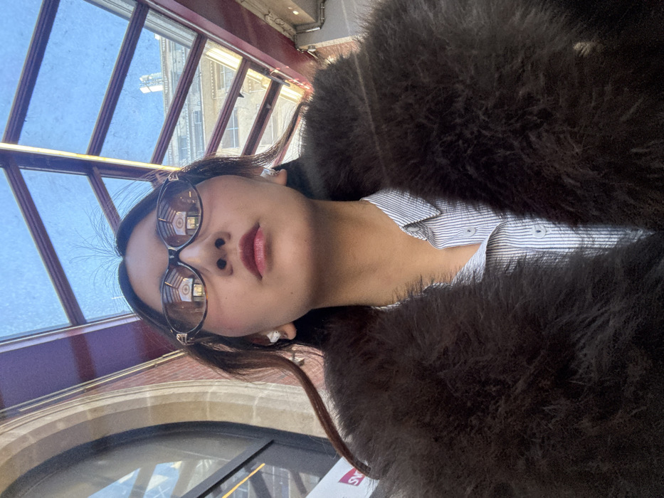 An individual is captured in a close-up, low-angle portrait, gazing slightly upwards. The person has long dark hair, wears dark tortoiseshell sunglasses with gold-colored detailing on the temples, and has white wireless earbuds in both ears. Red lipstick and flushed cheeks are visible. The individual is dressed in a dark brown fur coat or jacket over a light blue and white striped button-up shirt.
The background reveals a large, architecturally significant indoor space with ample natural light. On the left, a prominent arched window with light-colored stone framing is visible. Above, a multi-paneled glass ceiling or skylight structure, with dark framing, allows a clear blue sky to be seen. Reflections in the sunglasses show internal architectural elements, including what appears to be part of the vaulted ceiling structure. A white sign with the red "SNCF" logo is partially visible on the left. Through the glass roof, distant buildings exhibiting classic Parisian architectural styles are discernible. This scene is located within a concourse or hall of a major train station in Paris, France, identifiable as **Gare du Nord** due to the SNCF signage and characteristic 19th-century railway station architecture, including its distinctive glass and steel roof and stone detailing.