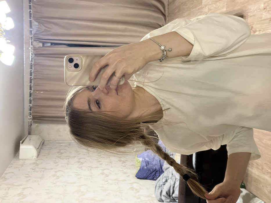 A light-haired female is depicted taking a mirror selfie in an indoor setting within the city of Zvezda, Russia. She wears a plain, light-colored long-sleeved top, a silver-toned charm bracelet on her right wrist, and a simple ring on her right ring finger. Her hair is styled in two braids, the left one of which she holds with her left hand. She holds a light-colored smartphone with a multi-lens camera in her right hand to capture the reflection. The background, visible in the mirror, includes light-patterned wallpaper, a white wall-mounted air conditioning unit on the upper left, and beige curtains on a white rod spanning the upper right. A portion of a ceiling light fixture with frosted shades is visible at the top. To the left, a dark piece of furniture, possibly a sofa or bed, is visible with a grey blanket and a dark blue pillow. The floor appears to be light-colored wood or laminate.