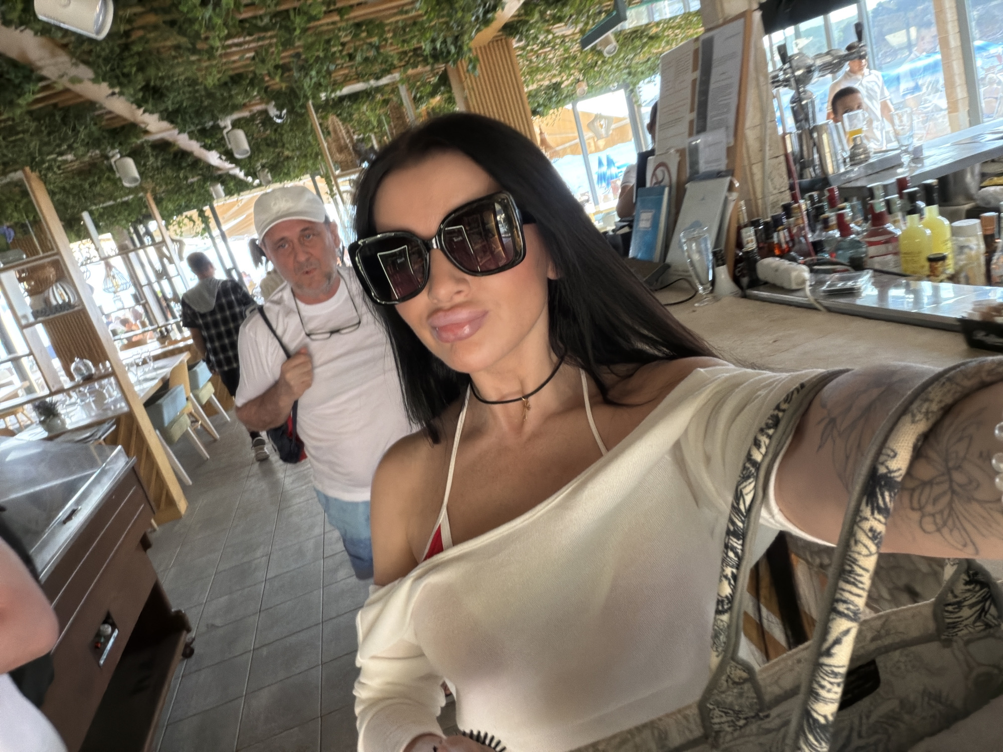 The image shows a woman taking a selfie in what appears to be a restaurant or bar, likely in Boreti, Montenegro. The woman has dark hair, sunglasses, and is wearing a white top with a bikini top underneath. A man in a white t-shirt and hat stands behind her to the left. Other patrons are visible in the background. The scene includes a bar area with liquor bottles and glasses, dining tables, and decorations. The environment suggests an open-air or beachside establishment, with glimpses of the outdoors visible through the windows.