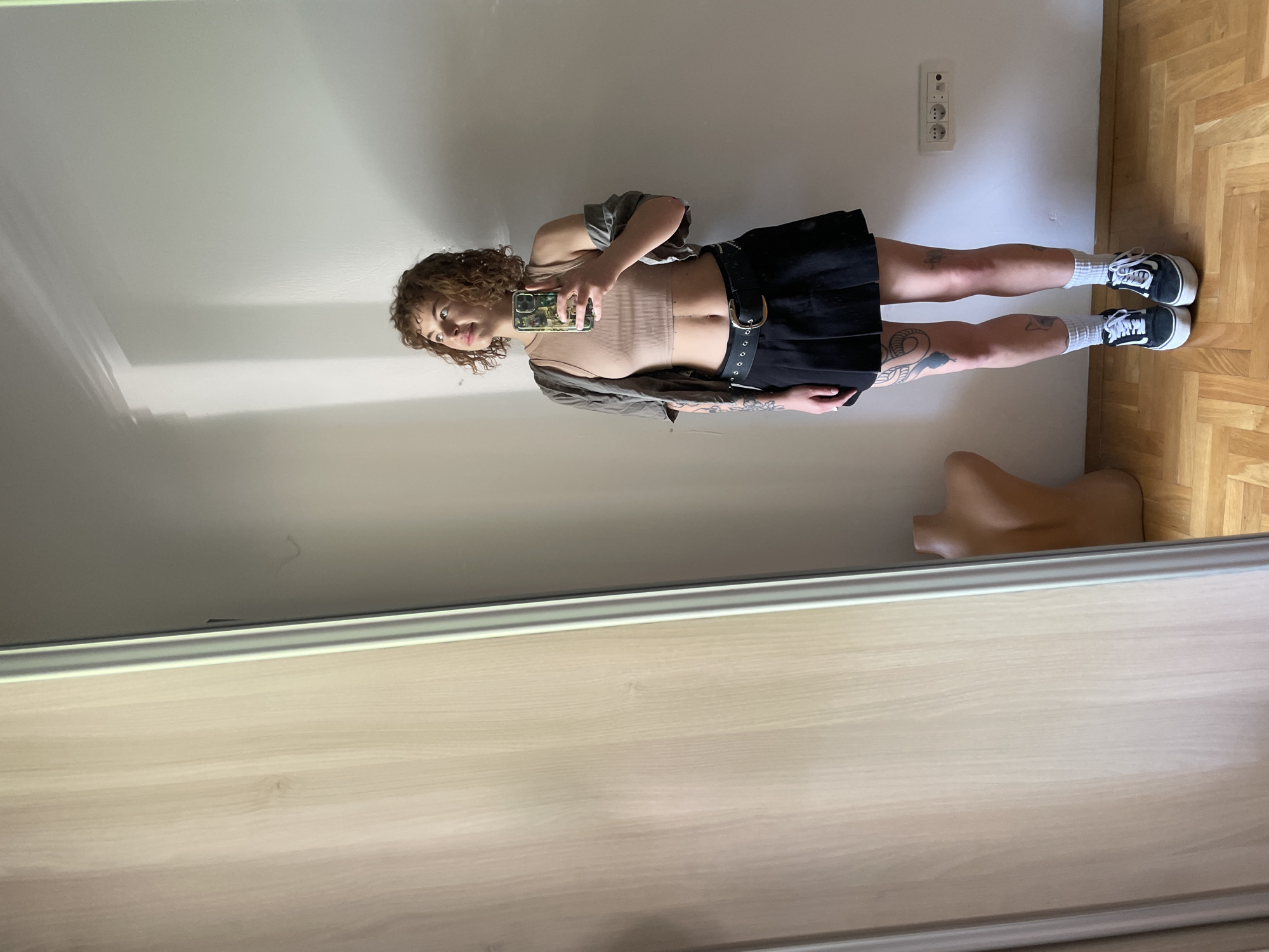 A woman stands indoors, reflected in a mirror. She is holding a phone in her right hand, presumably taking the picture. She has curly brown hair and is wearing a tan top, a grey shirt, a black skirt with a belt, white socks, and black and white sneakers. Tattoos are visible on her legs. The background includes a light-colored wall with an electrical outlet, a wooden floor, and what appears to be a mannequin leg. The location cannot be ascertained.