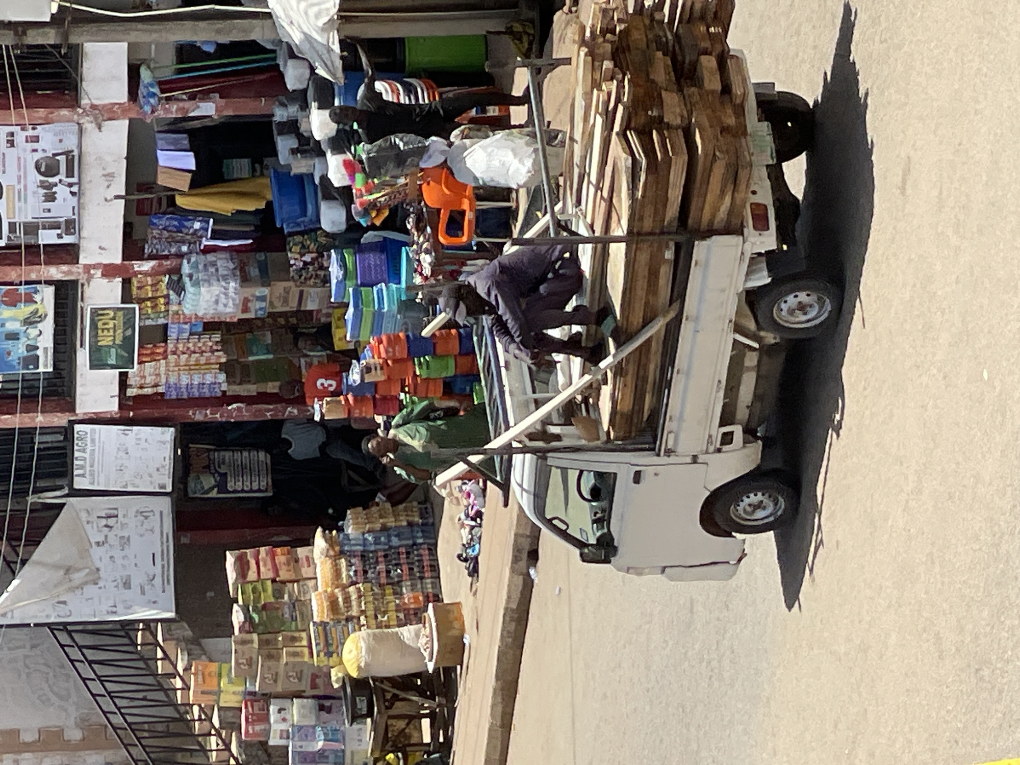 The image shows a bustling commercial scene in Bukuru, Nigeria. A small white truck is parked on a paved street, heavily loaded with planks of wood. A man wearing dark clothing is seen climbing on top of the load, presumably securing it. Next to the truck, a multi-story building serves as a market, displaying a wide array of goods, including clothing, plastic containers, and packaged foods. Storefront signs and advertisements are visible on the building's facade. Several people are present in the scene, some appear to be customers, while others could be vendors or passersby. The environment suggests a busy marketplace typical of an urban setting in Nigeria.
