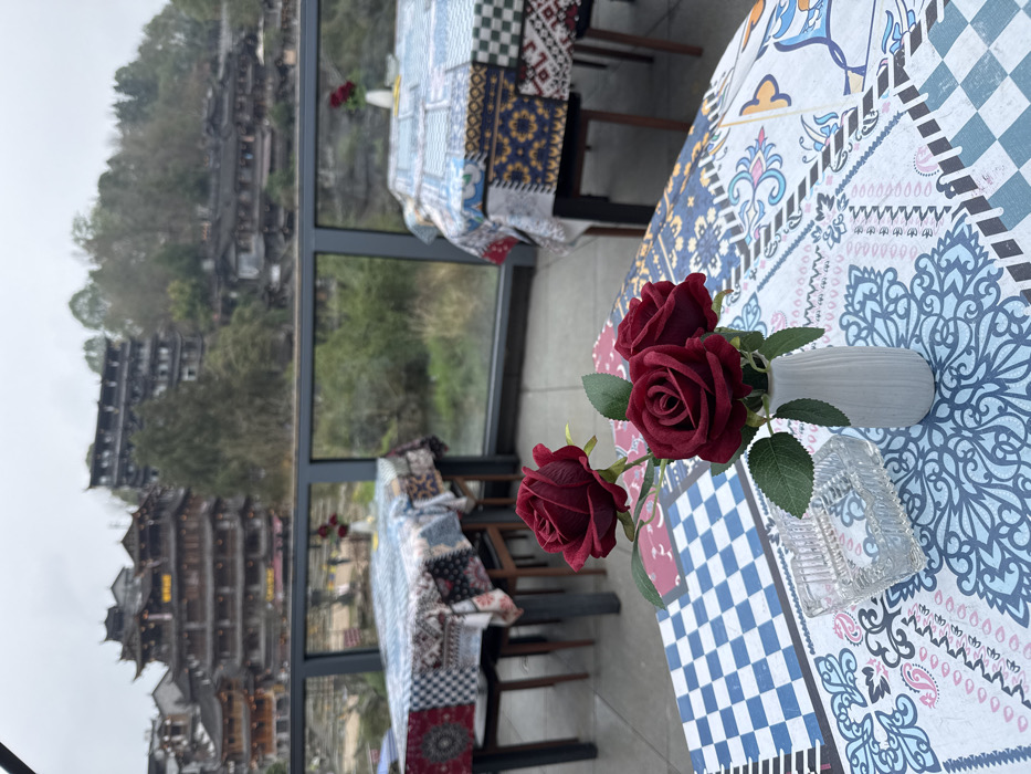 The image captures an outdoor dining patio in Furong, China, featuring a panoramic view of the ancient town under an overcast sky. In the foreground, a table covered with a patterned tablecloth displays a small white vase containing three artificial dark red roses and a clear, ribbed glass ashtray. Beyond this table, two additional tables are visible on the patio, each with a unique patchwork-style tablecloth and a small vase of red roses. A dark gray metal railing with clear glass panels defines the edge of the patio. Below the railing, a body of water or river is partially obscured by green foliage. The background showcases the historic city of Furong built into a steep, tree-covered hillside. Multiple multi-story buildings exhibiting traditional Chinese architecture, characterized by dark, tiered roofs and wooden facades, are prominent across the landscape. No individuals are discernible in the scene.