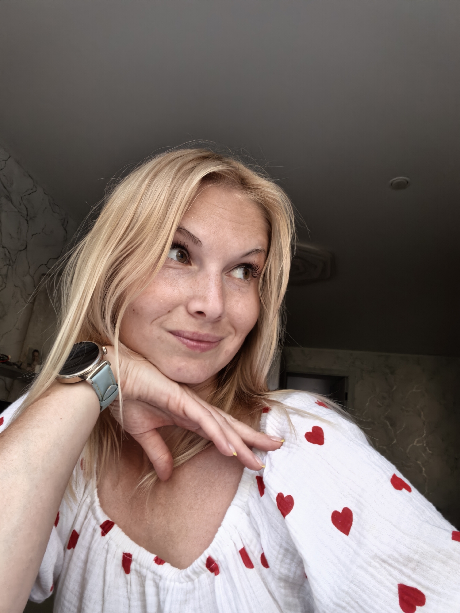 A blonde woman with light skin and freckles, wearing a white blouse with red heart patterns, rests her chin on her hand. She has a smart watch with a light blue band on her left wrist. Her eyes are looking to her right. The background appears to be an indoor setting with textured walls and a dark ceiling. There is no visible indication of the location being Sterlitamak, Russia.