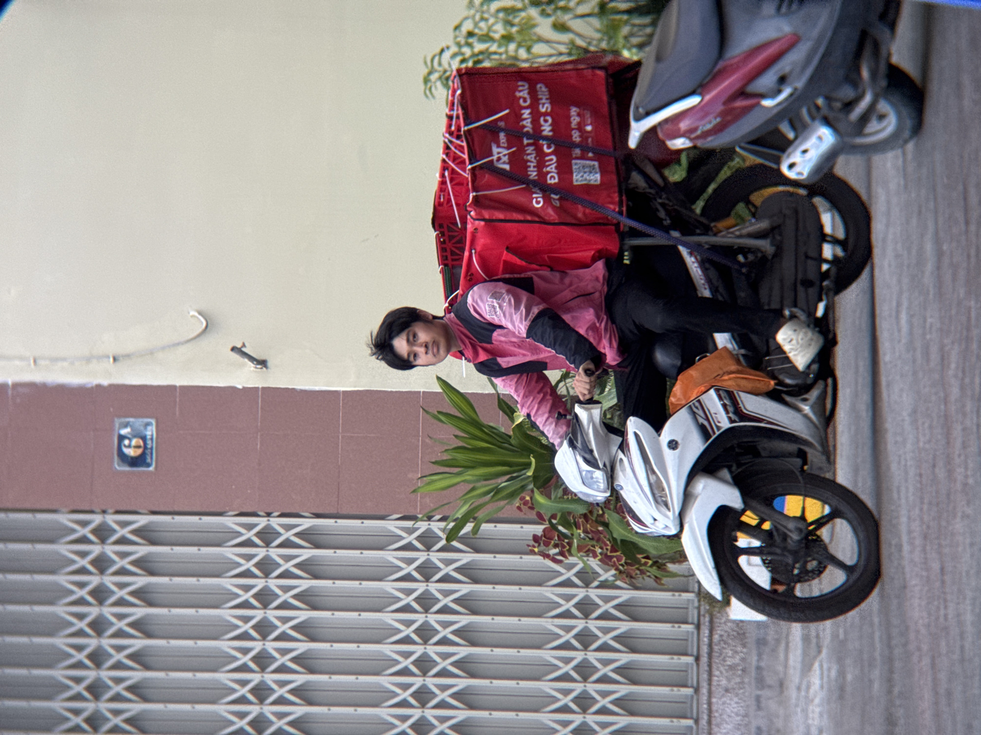 In Nha Trang, Vietnam, a young Vietnamese male delivery driver, wearing a pink and black jacket and black pants, is seated on a white scooter. He has dark hair and is looking forward. The scooter has a large red delivery box on the back, bearing Vietnamese text and a QR code. The driver's right leg is bent, and he is wearing white sneakers. An orange bag is attached to the side of the scooter. The scooter's front wheel is visible, and a Ukrainian flag is visible within the spokes of the wheel. To the right, a portion of a second scooter, dark red and grey, is visible. The background features a building with a textured brown and cream facade. A metal security gate with a crisscross pattern is to the left of the building, and a street number "6A" is mounted on the building. Green foliage is present between the scooters and the building. The ground is paved.