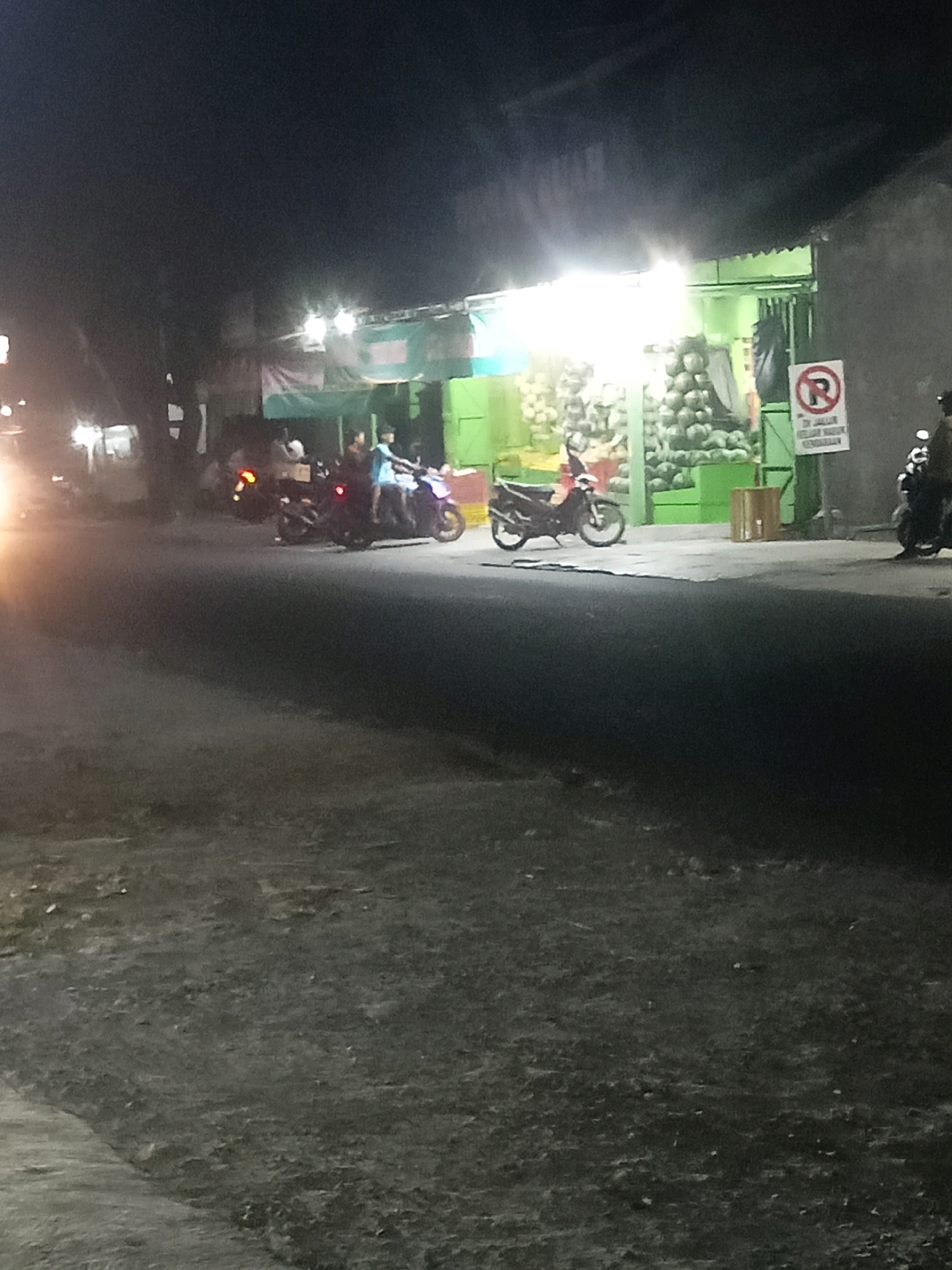 At night in Trihanggo, Indonesia, a busy street scene unfolds in front of a brightly lit building that appears to be a fruit stand. The building is painted green and stocked with what looks like a large quantity of coconuts. Several motorcycles are parked or in motion on the street, and people are visible interacting around the fruit stand and the vehicles. A "no parking" sign is prominently displayed on the right side of the building. The street itself is dark and appears to be paved, with a sidewalk or pathway in the foreground. The overall atmosphere is one of a lively, late-night market or roadside establishment.