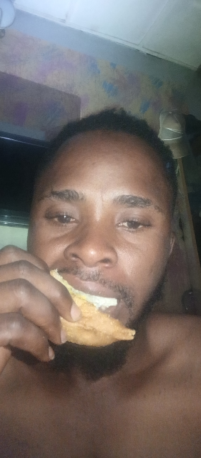 A close-up shot reveals a dark-skinned man with short, dark hair eating a pastry-like food. He is shirtless, his skin appearing damp. Behind him, a television screen and a wall with mottled, light-colored paint are visible. A white object hangs near the ceiling in the background. The focus is on the man’s face and the food he is holding.