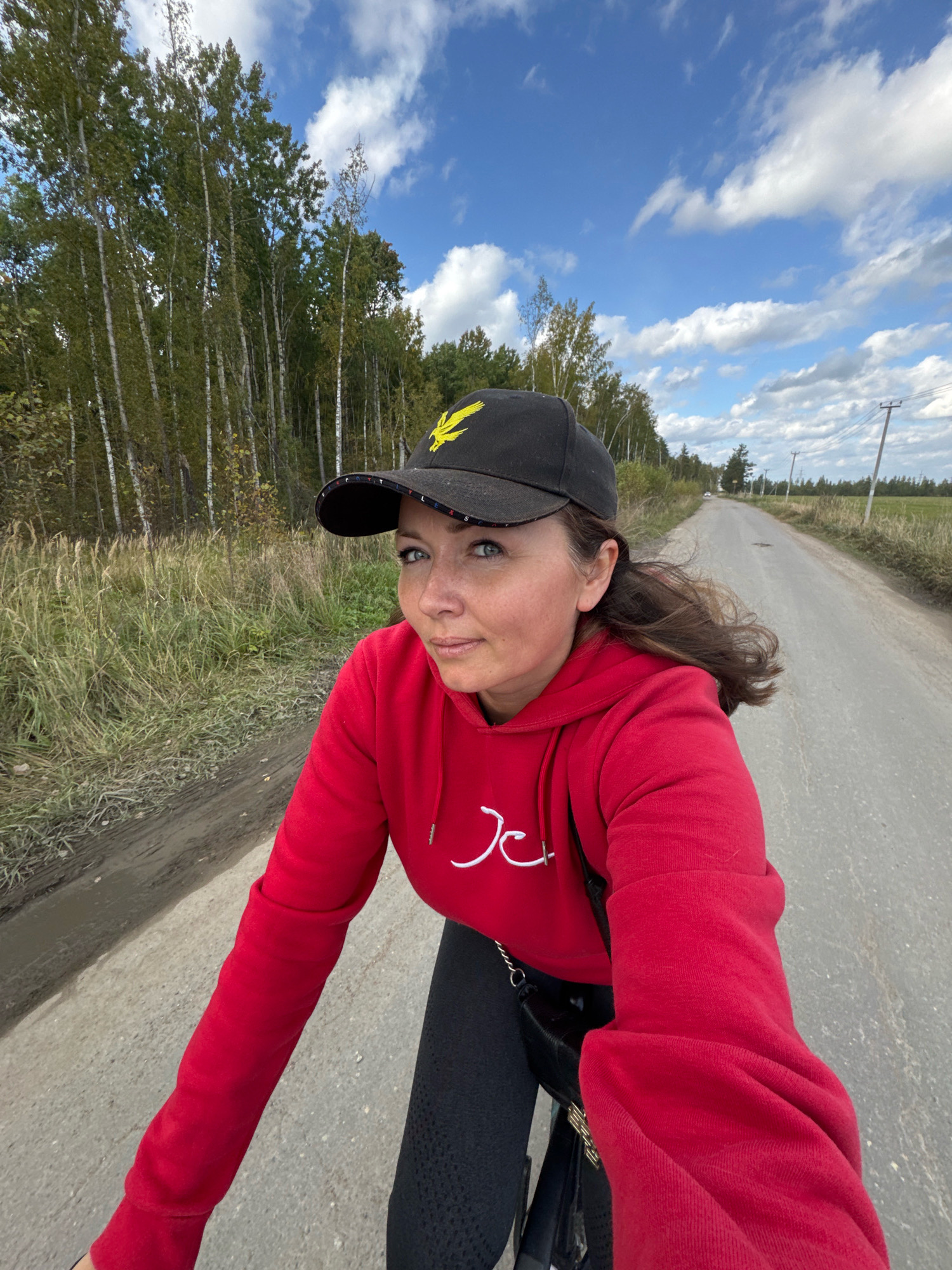 In a POV shot taken outdoors, a woman with brown hair, wearing a red hoodie and a black baseball cap with a yellow eagle emblem, is the central subject. She is looking towards the viewer with a neutral expression. Her left arm is extended, holding the camera. The background shows a dirt and gravel road, bordered by tall green grass and a dense forest of birch trees. The sky is blue with scattered white clouds, and there are telephone poles visible in the distance. The image was captured on a sunny day, with good lighting conditions. The location is identified as Innolovo, Russia.