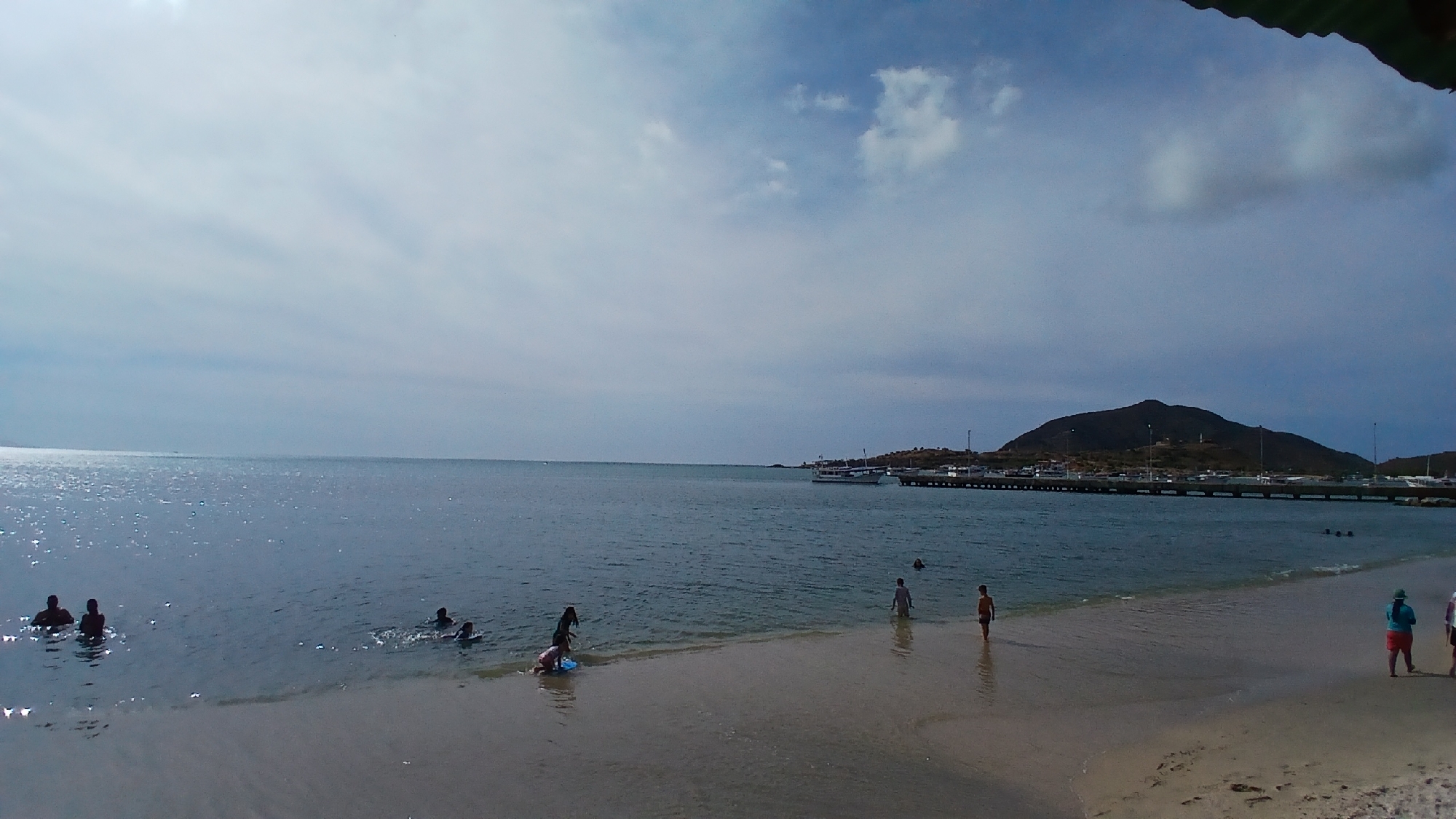 The image depicts a coastal scene in Juan Griego, Venezuela. The sky is partly cloudy with diffused sunlight reflecting on the water's surface. In the foreground, a sandy beach slopes into the ocean. Several people are in the water, some swimming or wading, while others are standing on the shore. In the background, there's a view of a harbor with boats docked along a pier. A landmass or hill is visible behind the harbor, shaping the coastline. The overall environment suggests a leisure or recreational setting.