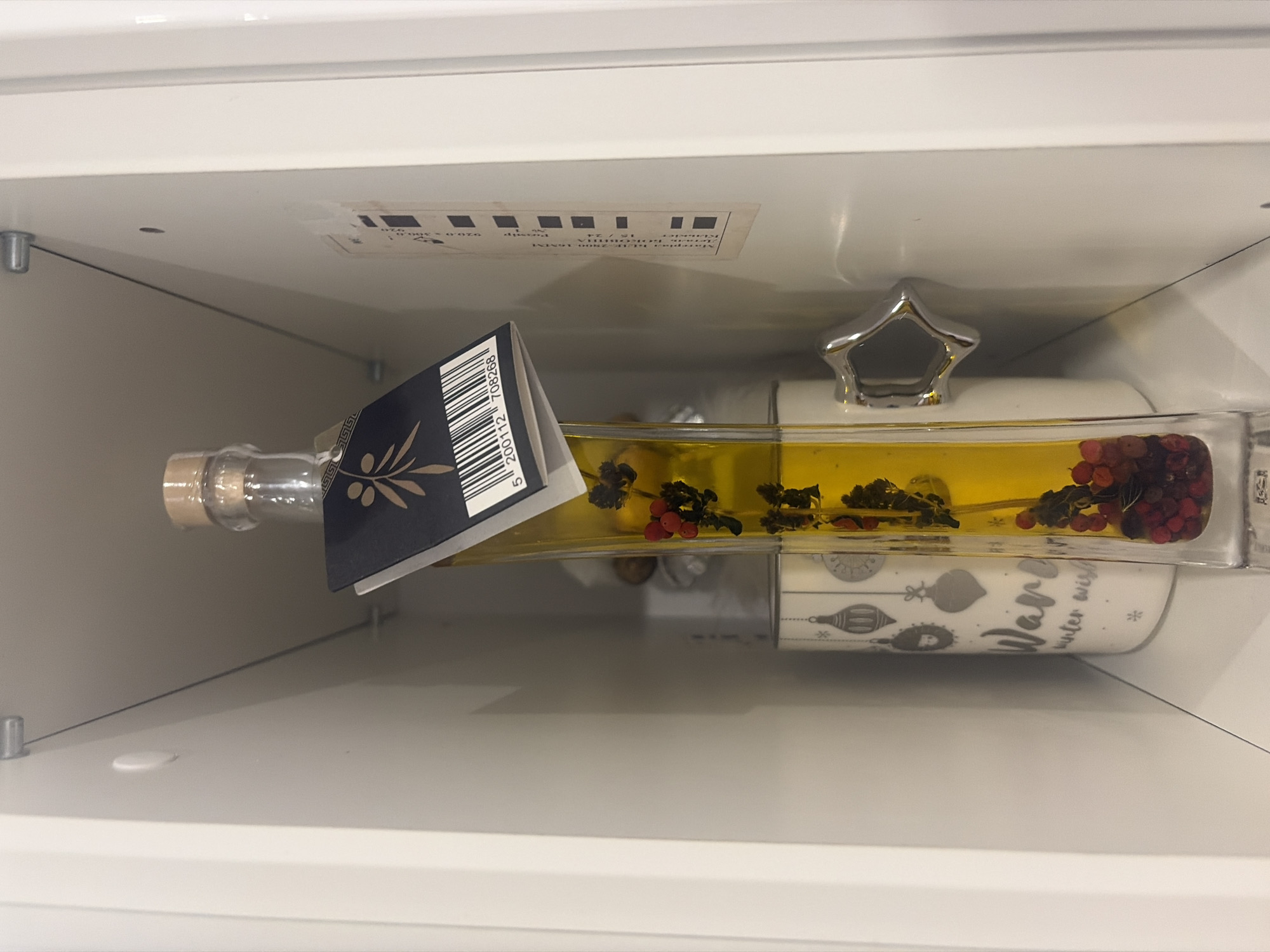 In a white, recessed shelf unit, a tall, clear glass bottle filled with golden oil and red berries is the central focus. A dark blue label with a golden olive branch design is attached to the neck of the bottle, featuring a barcode. To the left of the bottle, a white ceramic mug with a festive design of hanging ornaments and the text "Ware winter wish" is visible. A silver, star-shaped knob protrudes from the right side of the mug. The shelf unit has metal supports and a thin cable running diagonally across the back. The scene is indoors, likely a pantry or cupboard. The image was taken in Podgumer, Bulgaria.