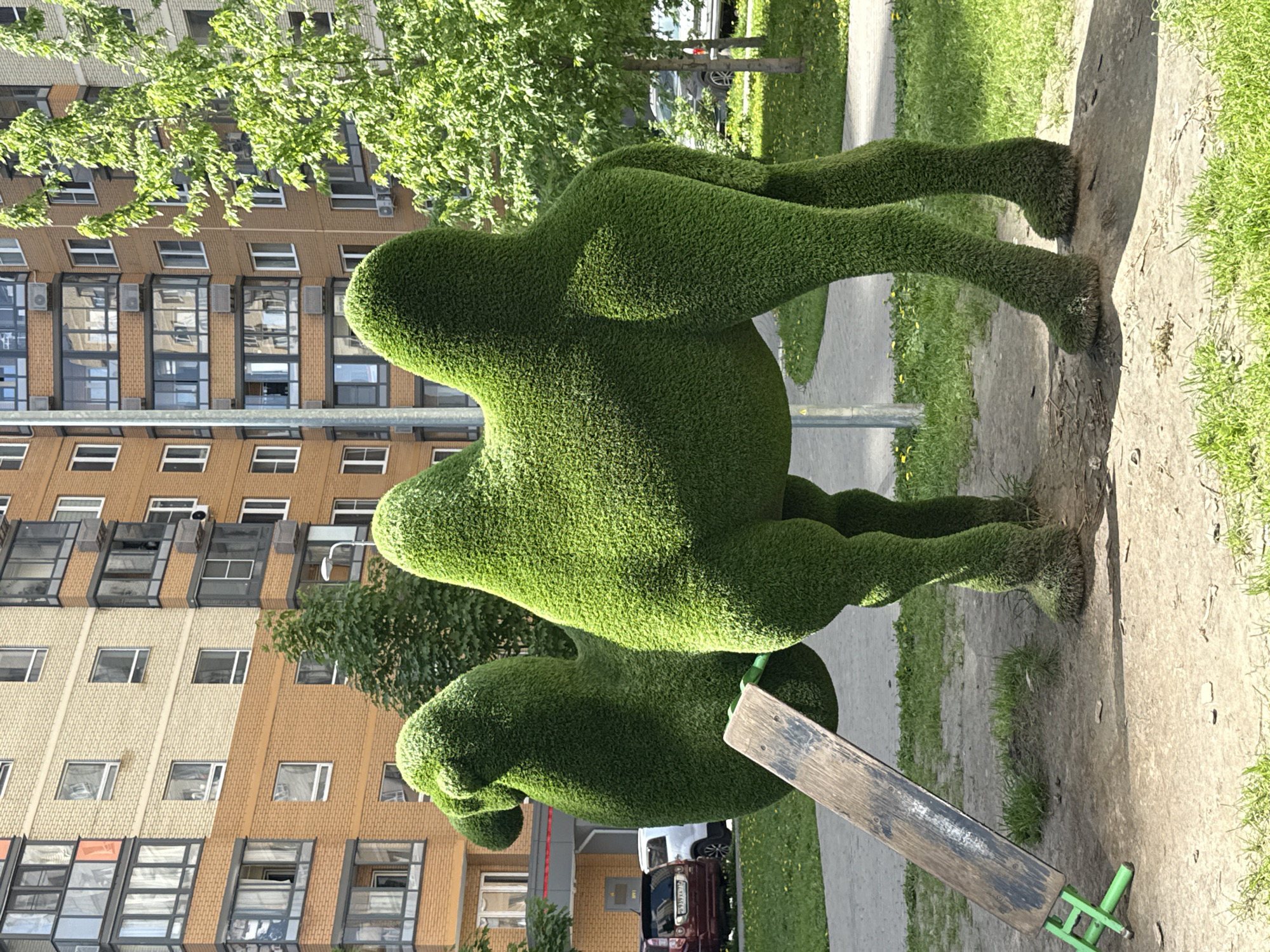 A green topiary of three people stacked on top of each other is on display. It is positioned next to a wooden and green metal seesaw on a concrete surface. A paved walkway and grass are to the side, and a multistory apartment building, trees, and vehicles are in the background. The location is in the city of Lyubertsy, Russia.