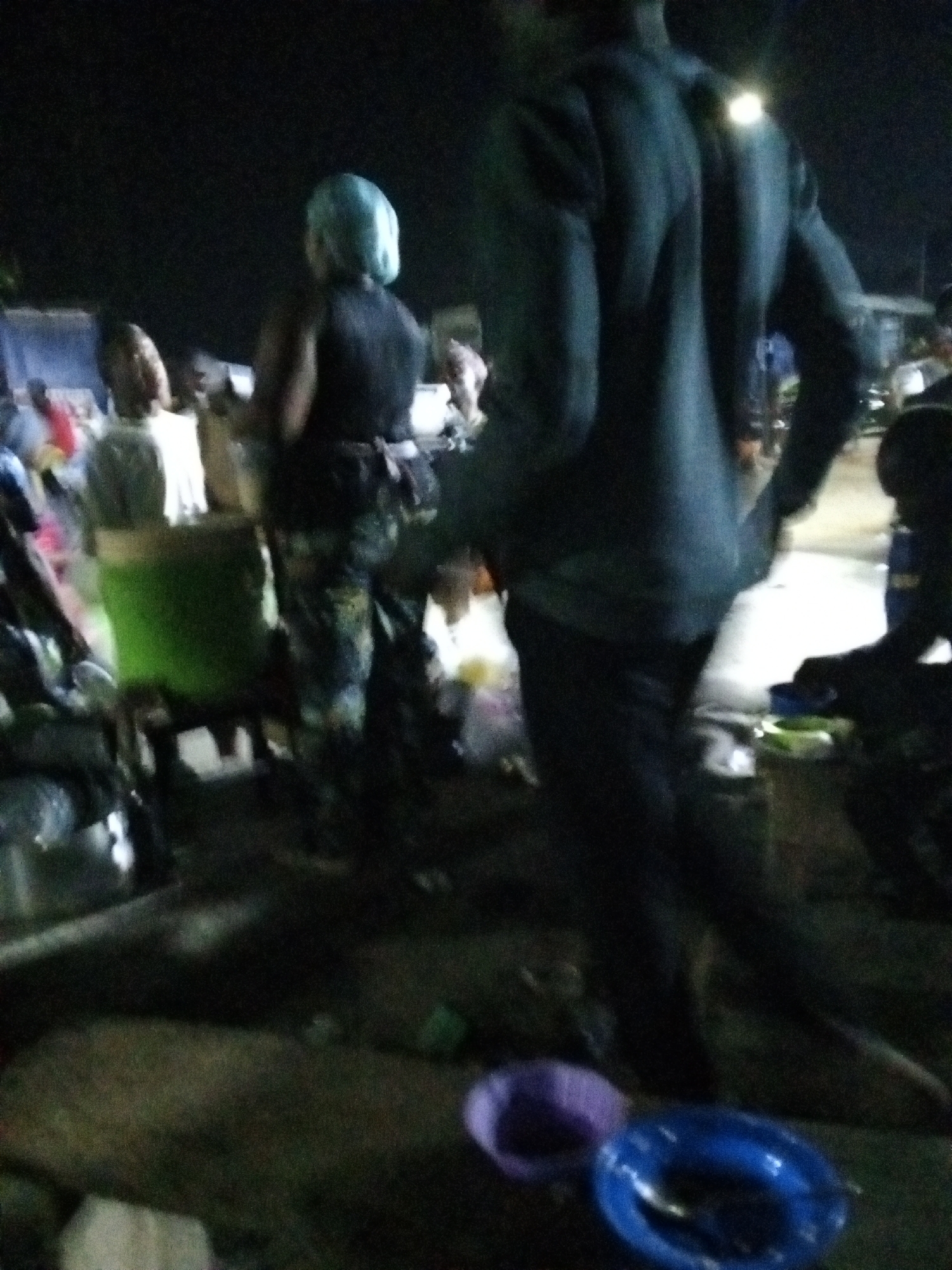 At night in an outdoor setting in Ado, Nigeria, a group of people are standing or walking amidst a poorly lit area with visible litter. A man in a dark suit stands in the foreground facing away from the camera. Near the bottom of the image are two plastic bowls, one purple and one blue, lying on the ground. Several other individuals are visible, including a woman with a blue head covering and patterned pants and a person standing behind a green object. There is a single street light illuminating the scene.