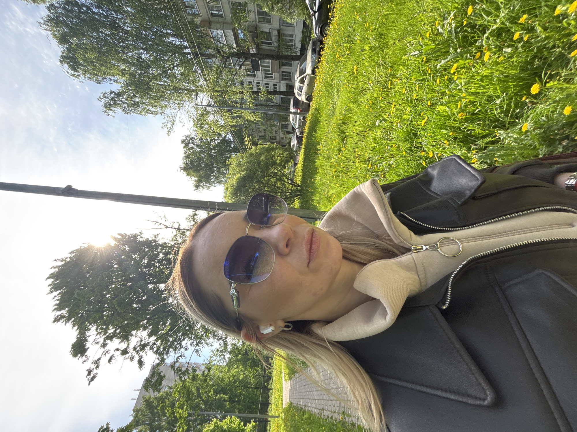 A selfie-style photo shows a woman in Saint Petersburg, Russia, on a sunny day. She wears sunglasses, a light-colored hoodie, and a black leather jacket. An earbud is visible in her ear. Behind her is a grassy area with yellow flowers and trees, with buildings and cars visible in the background. The overall scene conveys a relaxed, outdoor setting in an urban environment.