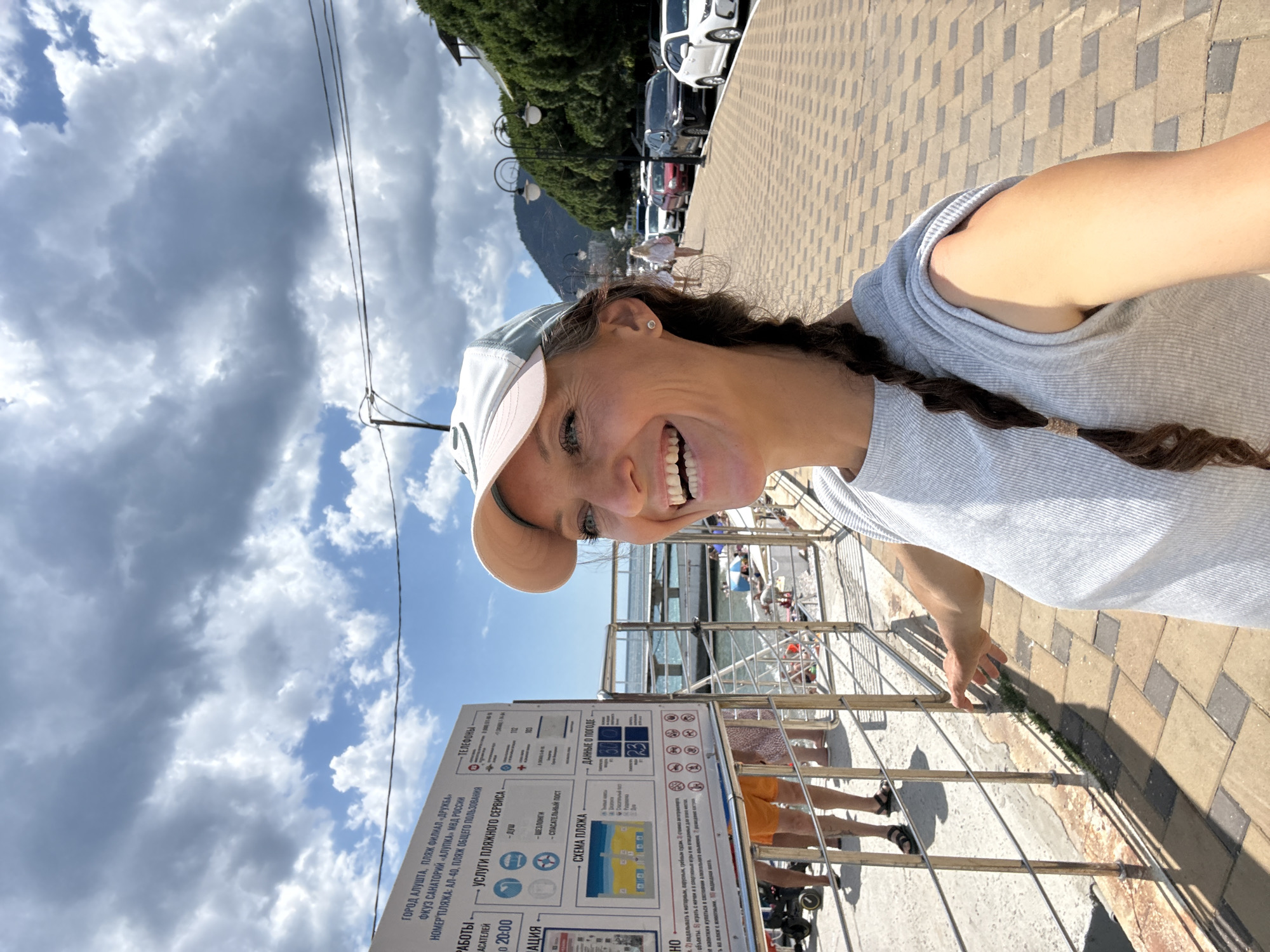 In the city of Alushta, Ukraine, a woman in a grey t-shirt and a white and pink baseball cap is taking a selfie on a sunny day. She is smiling broadly, with her hair in a braid. Behind her, a sign with Russian text provides information about the beach, including rules and services. A metal railing separates the boardwalk from the beach, where people are seen relaxing and swimming in the sea. Cars are parked along a tree-lined street in the background, with mountains visible in the distance. The sky is partly cloudy.