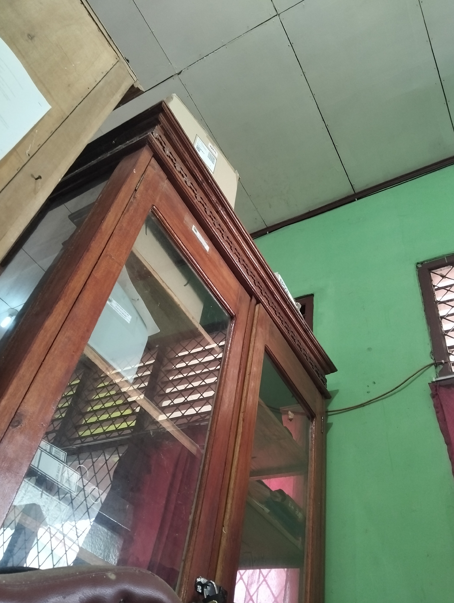 The image is an interior shot, likely from a residential setting in Banjar Agung, Indonesia. In the foreground, a wooden glass-fronted cabinet is prominent, with its top left corner angled towards the viewer. The cabinet appears to be made of dark, polished wood and features decorative carvings along its top edge. Through the glass doors, shelves are visible, some containing items that are not clearly discernible.
The background consists of a bright green wall. To the right of the cabinet, a window with dark wooden framing and a criss-cross pattern is visible. A dark red curtain hangs near the window. The ceiling, made of light-colored panels, is also visible in the upper portion of the image, showing seams and small nails. A light-colored wooden structure or shelf extends from the upper left corner of the frame, angled downwards. The overall lighting suggests an indoor environment, possibly during the daytime.