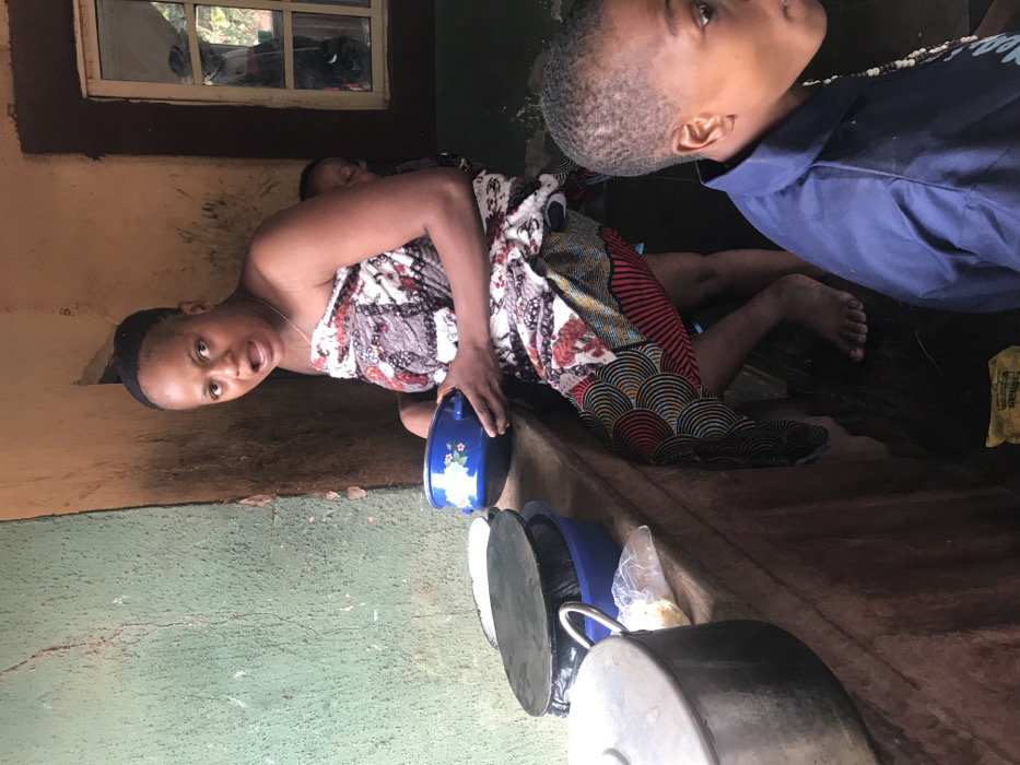This image depicts a domestic scene in Ogodo, Nigeria. The primary subject is an adult female, likely a mother, positioned centrally. She has a medium complexion, dark hair pulled back, and is wearing a multi-patterned fabric wrapper that covers her torso and lower body. Her mouth is open as if speaking or reacting. She is holding a blue enamel bowl with a white floral design in both hands. A baby is secured to her back with a plain fabric wrap, partially visible over her right shoulder. The woman stands barefoot on a dark, possibly wet or muddy, floor.
To her right and in the foreground, a young child with dark hair and complexion is partially visible, wearing a blue collared shirt. The child is looking towards the right side of the frame.
To the left of the woman, on an elevated, rough-surfaced, earthy-toned counter or ledge, several cooking utensils are visible. These include a large silver pot, two dark-colored pots or lids, and a blue plastic bowl. A clear plastic bag containing white granular material is also present on the counter. The wall behind the counter is painted a faded teal-green with visible cracks and wear.
The wall to the right of the woman is an earthy brown color, appearing rough-textured. A rectangular window with multiple panes, framed in a darker color, is set into this wall, offering a blurred view of an outdoor environment. The overall lighting suggests an indoor or semi-enclosed space, illuminated by natural light. A piece of yellow packaging material lies on the dark floor in the lower-right quadrant.
