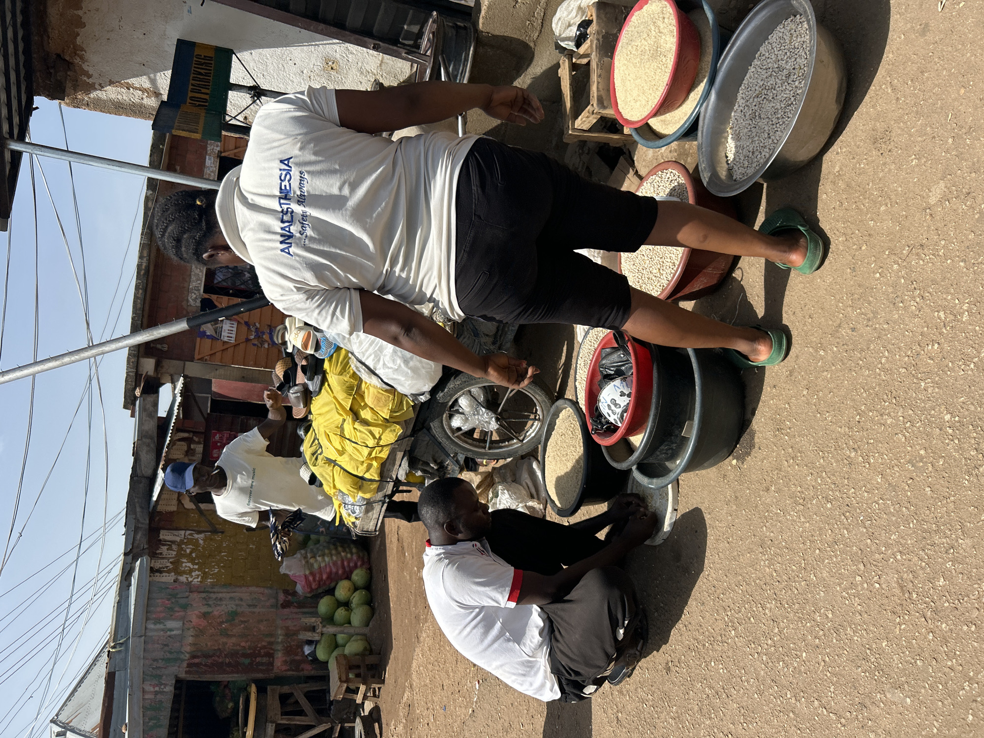 A daytime, medium shot captures an outdoor market scene with three individuals amidst sacks of grain and produce. In the foreground, a man wearing a white t-shirt with red trim and black pants is kneeling and sifting through a pile of grain on a white plate. To his right, a person in a white t-shirt with blue lettering that reads "ANAESTHESIA ...Safety Always" and black shorts stands facing away from the camera, looking towards the market stalls. In the background, a man in a white t-shirt and a blue baseball cap is standing near a cart laden with yellow sacks, holding what appears to be a cigarette and a striped cloth. The environment is dusty with a dirt ground, and in the background, there are simple market structures constructed from corrugated metal and wood, with overhead wires crisscrossing the sky. The scene suggests a commercial transaction or preparation of goods for sale.