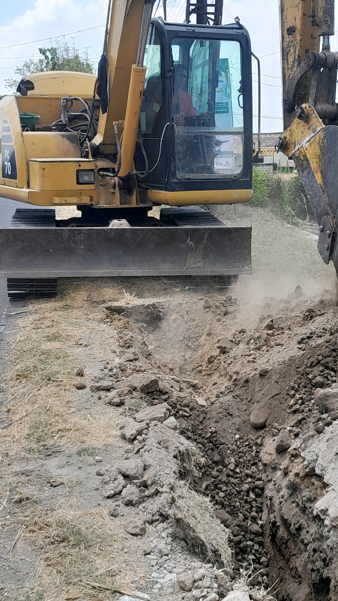 In Jomblang, Indonesia, an excavator is actively engaged in excavation work on a roadside. The excavator, a yellow Caterpillar PC70 model, is positioned on the left side of the frame, its tracks resting on the ground. A person, wearing an orange shirt, is visible inside the excavator's cabin, operating the machinery. In front of the excavator, a blade is lowered, pushing dirt and debris. To the right of the excavator, a trench is being dug, with loose soil, rocks, and dry grass scattered around the excavation site. The background shows a mix of vegetation and some structures, suggesting an outdoor, possibly rural or semi-urban environment. The overall scene depicts an ongoing construction or roadwork activity.