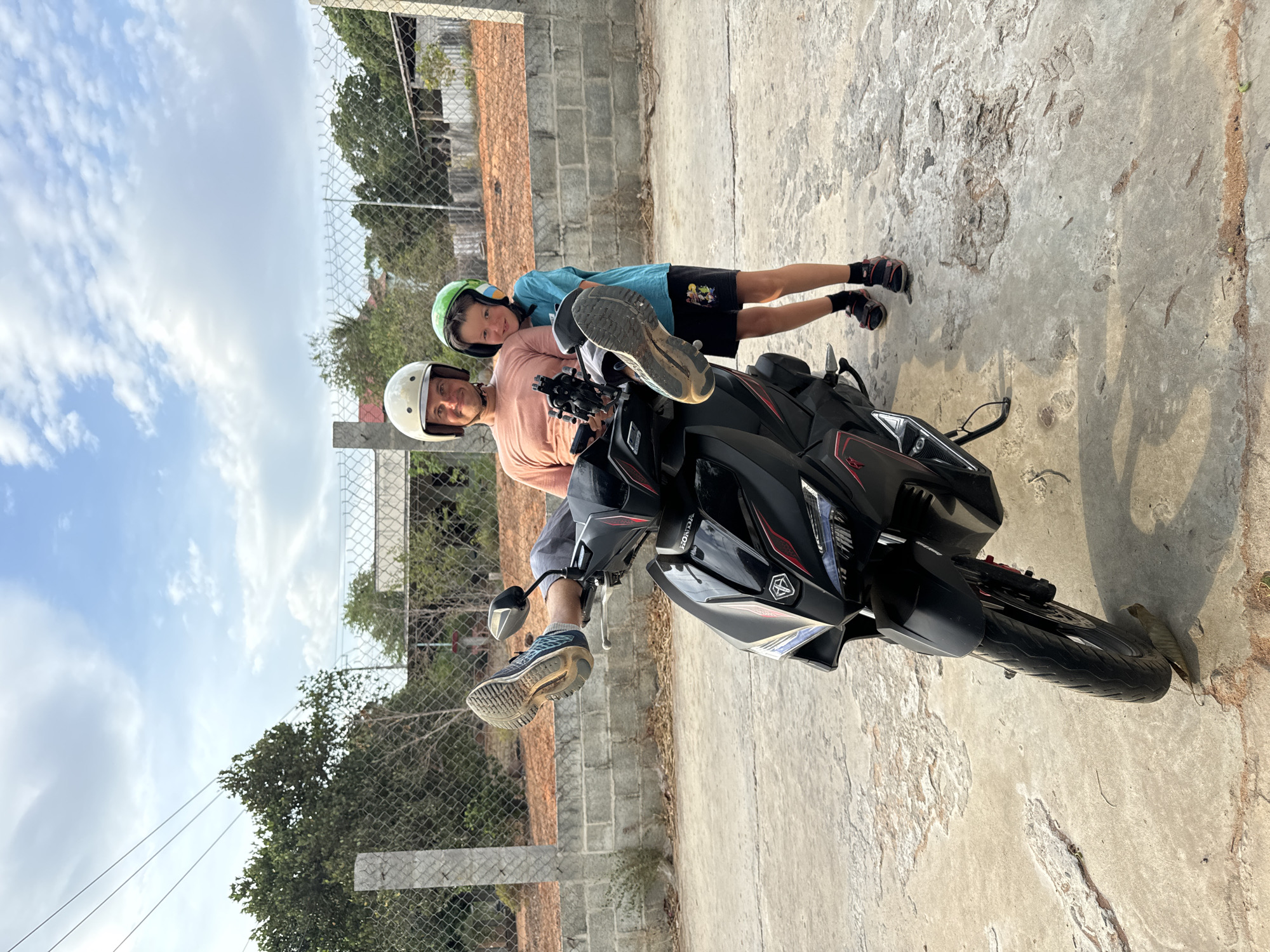 Two people are straddling a motorcycle. One person in a peach shirt and white helmet is sitting on the motorcycle with their legs extended. Another person in a blue shirt and green helmet is lying prone over the person in the peach shirt with their legs extended behind the motorcycle. The motorcycle is black, and the photo is taken outside on a concrete surface. There is a fence behind the people and vegetation behind the fence. The location is identified as Mũi Né, Vietnam.