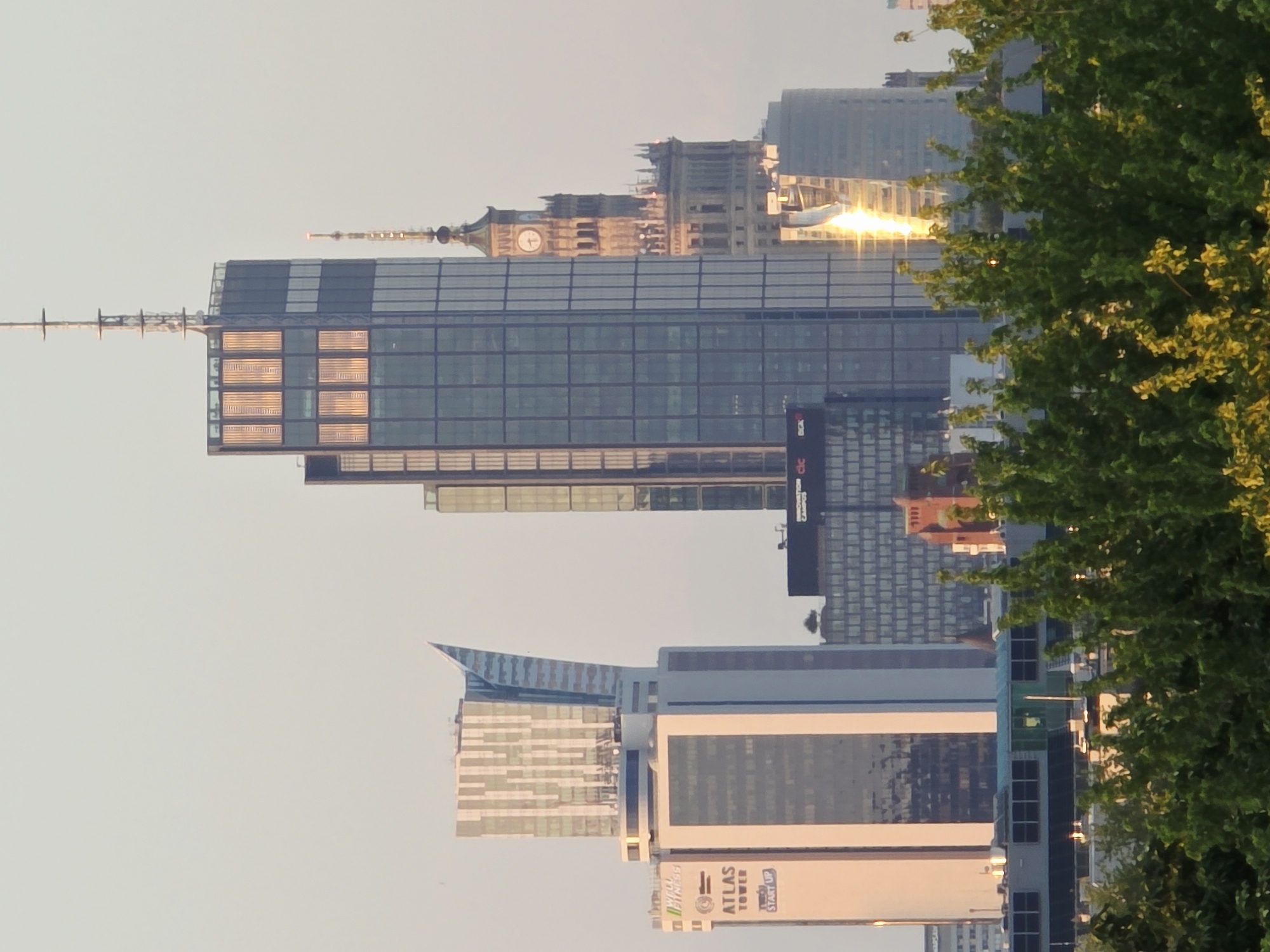 The image shows a cityscape of Warsaw, Poland, featuring several prominent skyscrapers. In the foreground, lush green trees obscure the lower part of the scene. The main focus is on a collection of modern and older architectural styles. To the left, a lighter colored building with a unique angular top is visible, marked with the text "ATLAS TOWER". Adjacent to it and slightly behind is a taller, darker skyscraper with numerous windows, reflecting the golden light of the setting sun. To the right of this, a more ornate, older tower with a clock face stands out. Further in the background, other buildings of varying heights and designs are present, contributing to the urban skyline. The sky is a pale, clear hue, suggesting late afternoon or early evening.