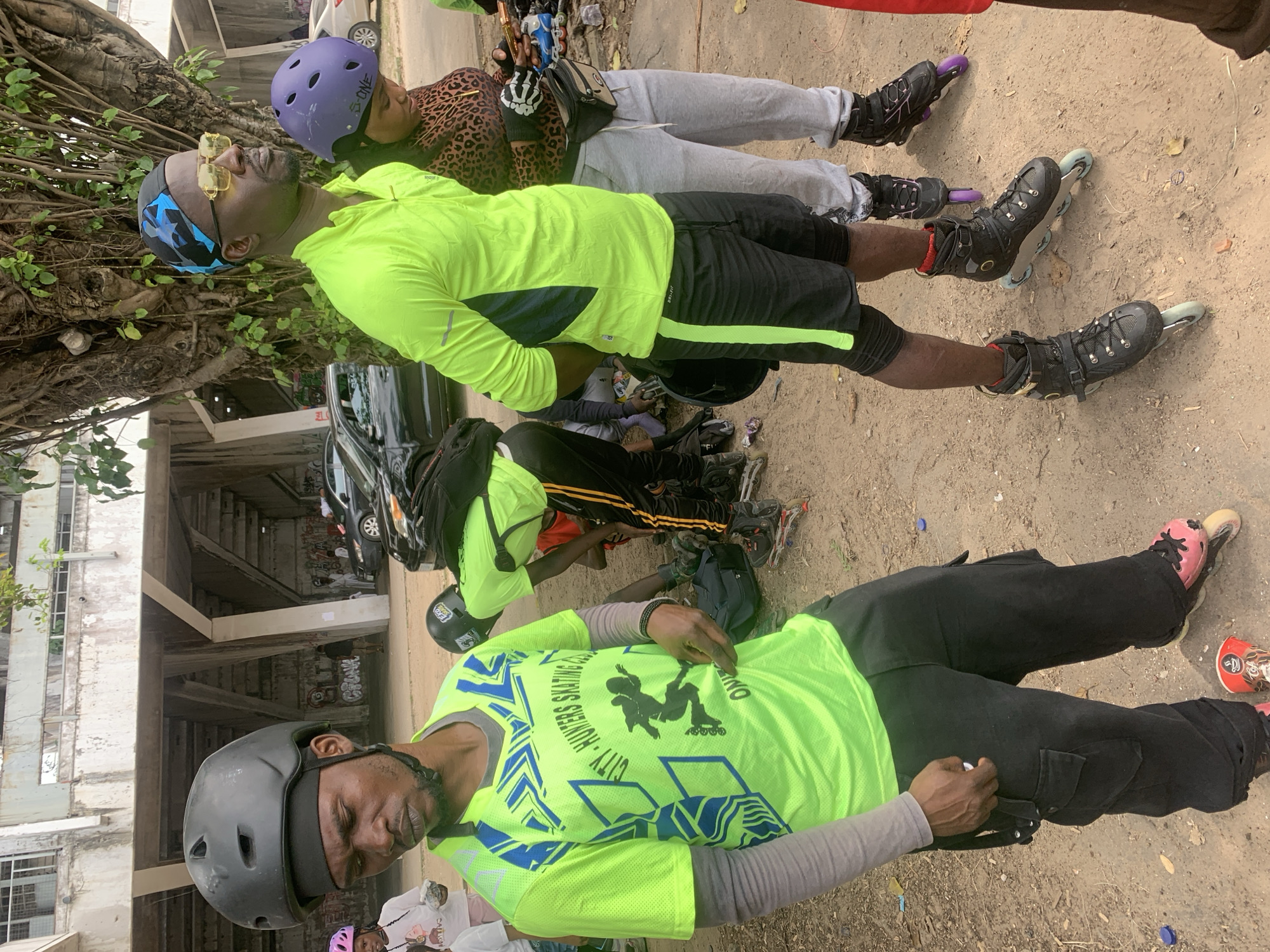 In Alagbede, Nigeria, a group of people are gathered outdoors near a concrete structure with tiered seating, possibly a stadium or amphitheater. A large tree with dense foliage dominates the background. In the foreground, a man wearing a black helmet and a neon yellow long-sleeved shirt with blue graphics is the central figure. His shirt displays the text "CITY - HUNTERS SKATING CLUB" and an image of a skater. He is also wearing black cargo pants and rollerblades. To his right, a man with sunglasses and a blue and black bandana is wearing a neon yellow long-sleeved shirt and black shorts. Next to him, a woman with a purple helmet and a leopard-print top is standing with grey sweatpants. Several other individuals, some wearing helmets and rollerblades, are visible in the background, along with cars and scattered debris on the ground. The scene appears to be a gathering for roller skating enthusiasts.