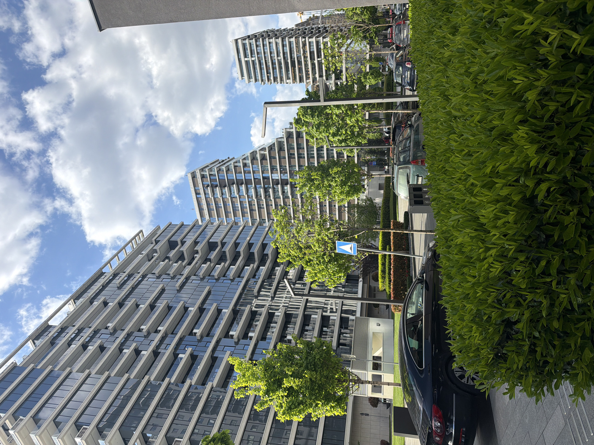 This is a daytime photograph depicting a modern urban street in Belgrade, Serbia. The scene is dominated by contemporary high-rise buildings on the left, an arterial road or wide pavement with parked cars on the right, and a bright blue sky with white clouds at the top.
The buildings, primarily on the left side of the frame, are multi-story structures featuring sleek, modern architecture with extensive glass facades, angular concrete elements, and numerous balconies or recessed areas. They suggest a newly developed residential or mixed-use complex.
To the right, a well-maintained street or large paved area is visible, bordered by a dense, manicured green hedge that runs along the right edge of the frame. Several trees with vibrant green foliage are planted along the thoroughfare, casting light shadows.
A line of cars is parked along the street, partially obscured by the hedge. A dark-colored vehicle is prominent in the mid-ground, with its side and rear visible. Another vehicle's license plate shows the numbers '2299'. A blue triangular sign with a white arrow pointing upwards is visible between two trees on the street.
The clear blue sky with fluffy white cumulus clouds suggests sunny and pleasant weather conditions, likely mid-morning or early afternoon given the bright illumination. The overall impression is one of a clean, modern, and well-organized urban environment.
