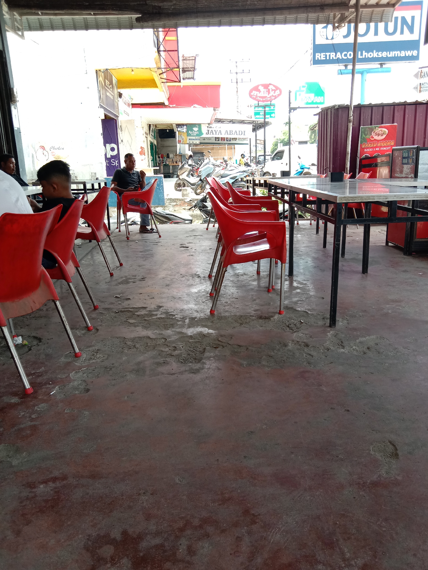 The image shows an outdoor dining area, presumably in Lhokseumawe, Indonesia, based on the signage visible in the background. There are several red plastic chairs and tables with metal frames arranged on a concrete floor. In the background, people are seated, and several motorbikes are parked outside businesses like Avian and Jaya Abadi. Storefront signs include "Retraco Lhokseumawe," "Malika Bakery and Cake Shop," and a promotional sign for "Bakso Aroma." The scene is partly covered by a corrugated roof, providing shade to the dining area.