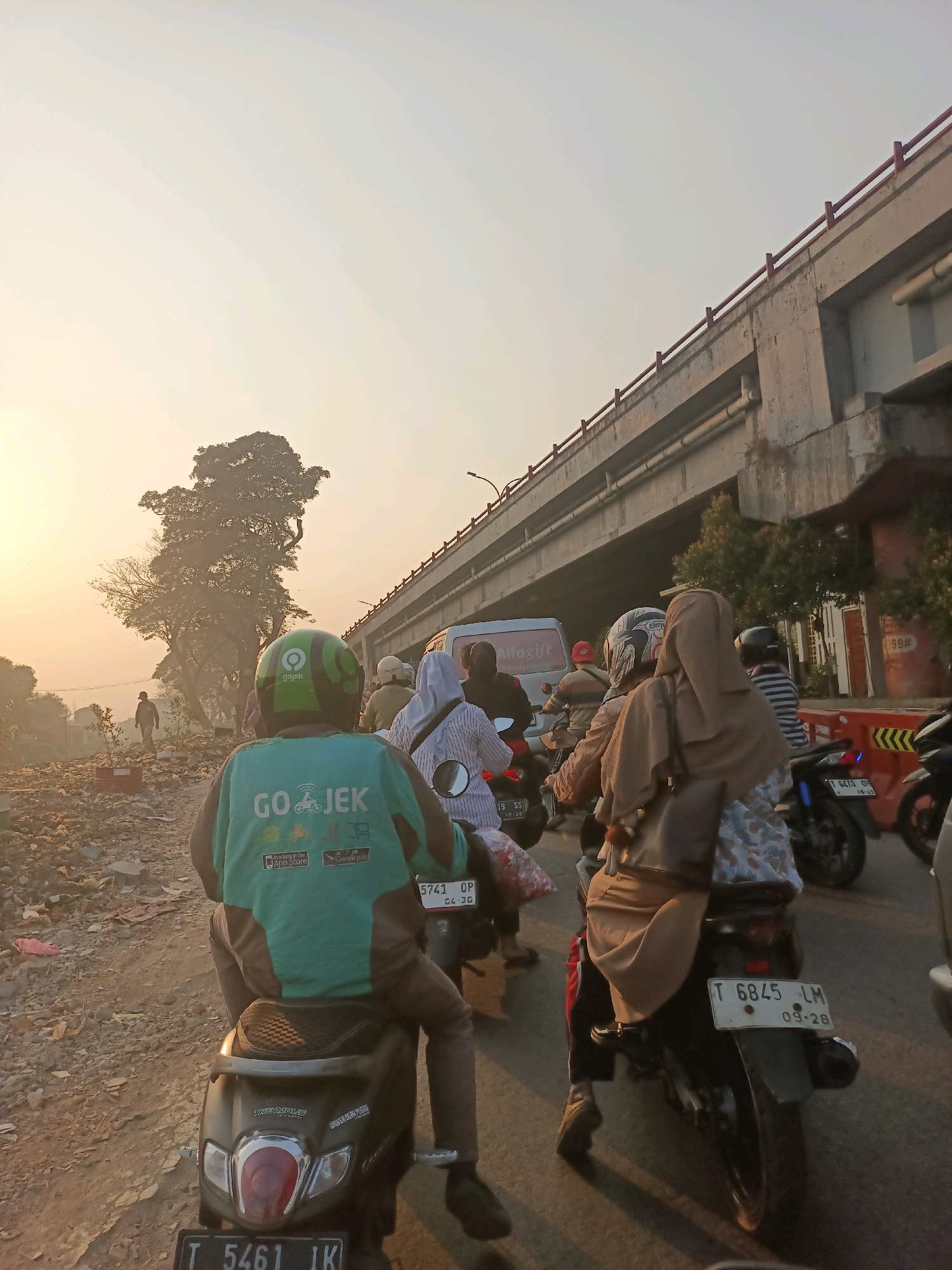 The image depicts a street scene in Karawang, Indonesia, during what appears to be either sunrise or sunset, given the warm, hazy lighting. The foreground is dominated by a person riding a motorcycle, wearing a green Gojek helmet and a teal jacket with the Gojek logo. The motorcycle has a license plate visible, reading "T 5461 IK".
Behind this motorcycle, a series of other motorcycles and vehicles are visible, suggesting traffic congestion. Many of the riders and passengers are wearing headscarves, indicating a likely female presence among them. The road surface appears to be a mix of asphalt and dirt, with debris scattered along the sides.
In the background, a multi-level overpass or bridge structure is prominent, with concrete supports and a red railing along the top. Trees are visible near the overpass, some appearing bare or with sparse foliage. A white van with "Alfogift" branding is also present in the traffic. The overall environment suggests an urban or semi-urban setting in Indonesia.