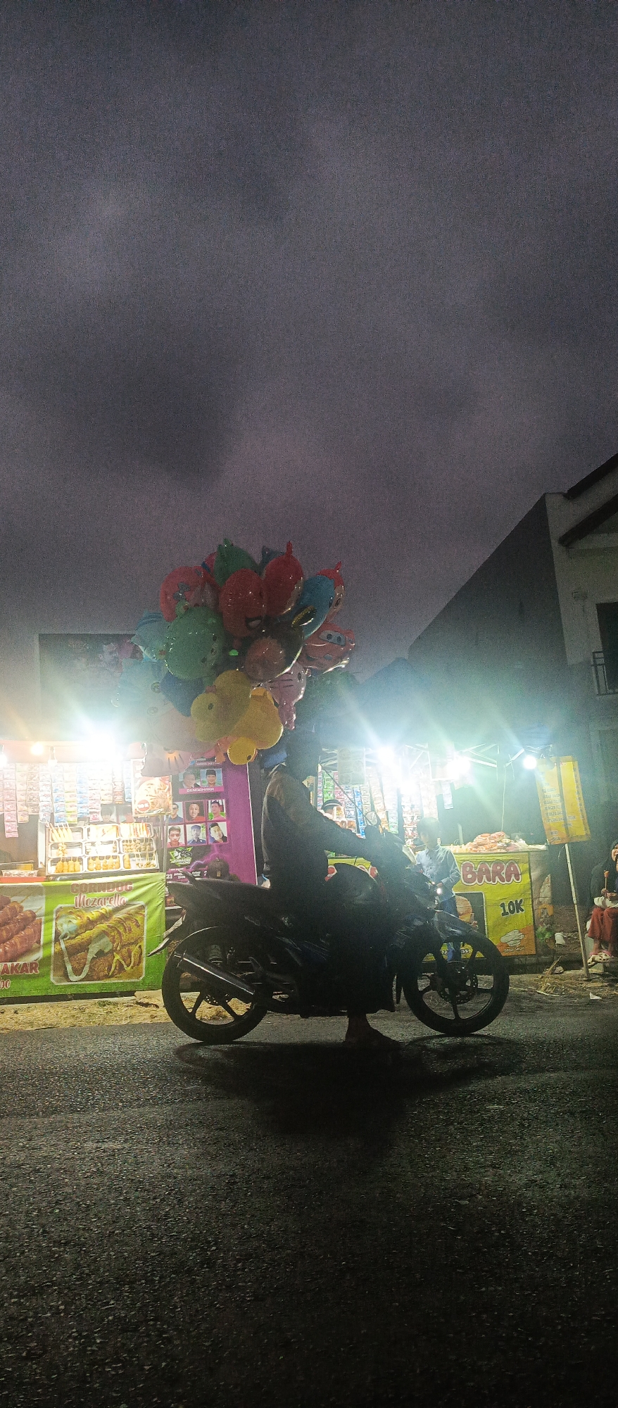 A man on a motorcycle carrying a large bunch of colorful, character-shaped balloons is parked on a street in Sumbersekar, Indonesia. The scene takes place at dusk or night, with bright artificial lights illuminating various market stalls. In the background, there are food vendors selling items like corn dogs and snacks. The sky is dark and overcast. The street is asphalt.