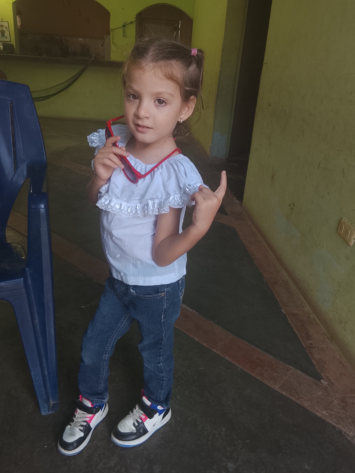 A young girl stands indoors, possibly in a home or public space in Altagracia, Venezuela. She is wearing blue jeans, a white blouse with lace trim, and multi-colored sneakers (predominantly black, white, and pink). In her left hand, she holds red sunglasses. Her hair is pulled back into pigtails. She is facing forward, looking at the camera, and making a hand gesture with her right hand. To her left is a blue plastic chair. The wall behind her is yellow and appears to be somewhat aged. In the background, through an archway, there is what appears to be a hammock hanging and a table with items on it. The flooring has a dark, textured pattern, with visible seams and cracks. There's an electrical outlet visible on the wall behind her.