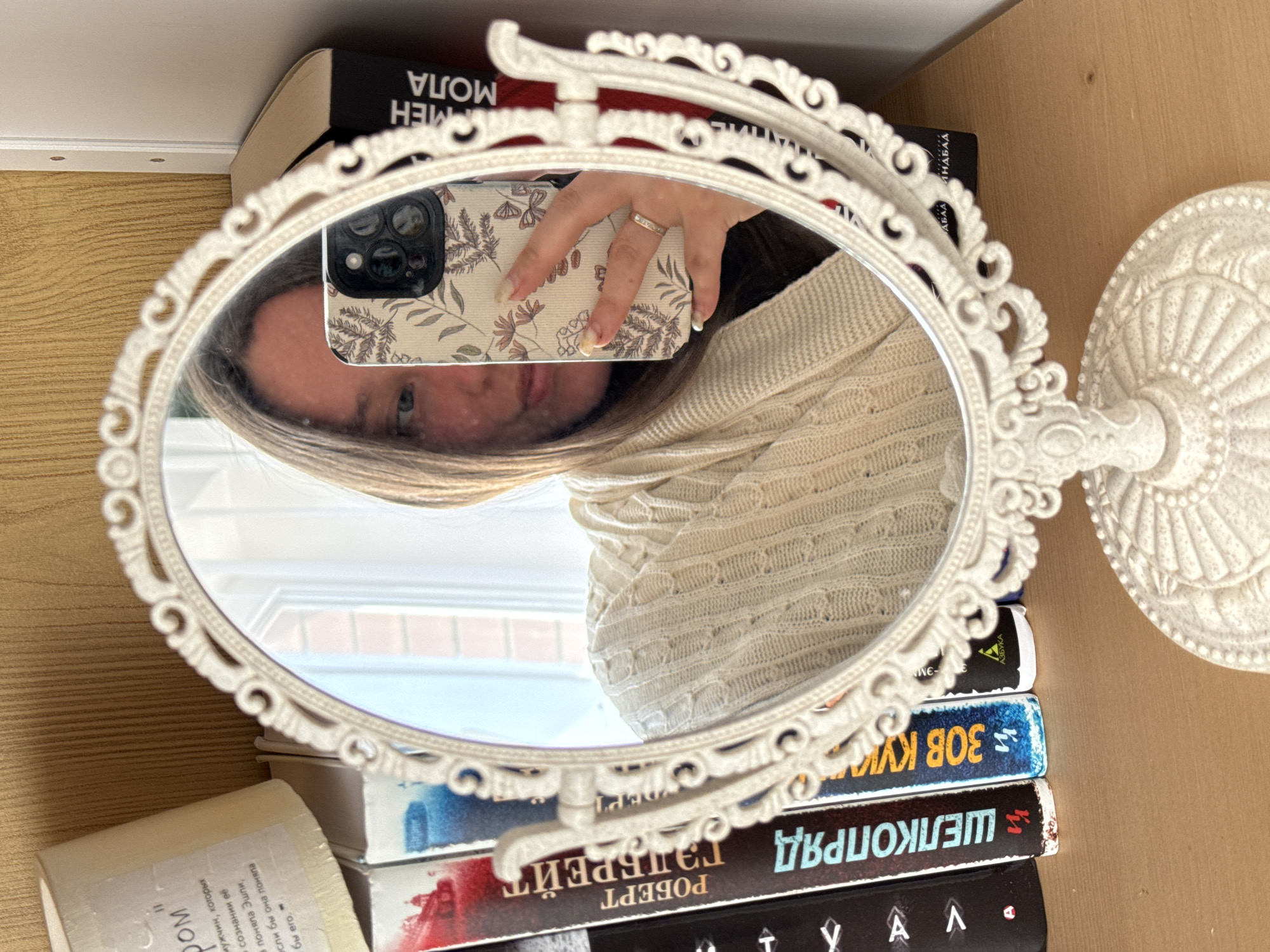 The image displays an indoor mirror selfie, likely taken in Krasnodar, Russia, during daytime hours due to the apparent natural light. The main subject is an ornate, oval-shaped white mirror featuring a decorative, antique-style frame and stand.
Reflected in the mirror is a person, likely female, with long, light brown hair, wearing a light-colored, possibly cream or beige, cable-knit sweater. Only a portion of their face, including one eye, is visible. The person is holding a smartphone with a distinctive cream-colored case decorated with a pattern of leaves and butterflies or moths. A ring is visible on the ring finger of the hand holding the phone.
The mirror and its stand are positioned on a stack of several books, which rest on a wooden surface, possibly a shelf or desk. The books feature Cyrillic script, indicating they are in Russian. Visible text includes the titles "ШЕЛКОПРЯД ГЭЛБРЕЙТЕ" (The Silkworm by Robert Galbraith) and "ЗОВ КУК" (likely part of "The Cuckoo's Calling" by Robert Galbraith), along with the author's name "РОБЕРТ". Other snippets of text include "АЗБУК" (likely from the publisher Azbuka), "МОЛА", and a phrase that translates to "men, whom Ashley understood in her mind, if she understood him," possibly a book description. The spine of the bottom book shows "А Λ" and "D." The overall setting suggests a personal and cozy space.