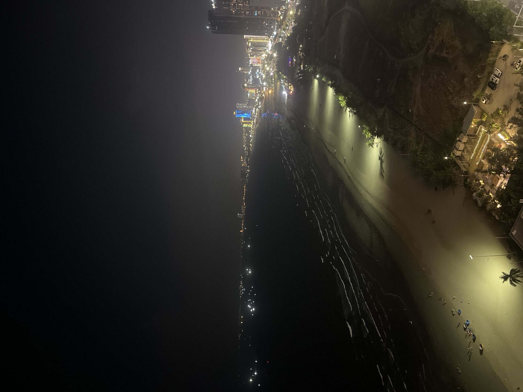 This is an aerial nighttime shot of a beach in Đà Nẵng, Vietnam. The image is rotated 90 degrees counterclockwise. The scene captures a lively beachfront with a city skyline in the background.
The left side of the image is dominated by the dark ocean, with the faint glow of distant lights reflecting on its surface. The beach itself is visible on the right, stretching along the coastline. It appears to be a sandy beach, with some areas of dark, wet sand closer to the water, indicating the tide has recently receded.
Along the beach, there are scattered lights from streetlamps, illuminating sections of the sand. Several small groups of people are visible on the beach, appearing as tiny figures enjoying the evening. Some are walking, while others seem to be gathered near the water's edge. There are also several colorful objects, likely beach towels or chairs, scattered on the sand.
Further inland, lining the beach, is a strip of palm trees and vegetation, also illuminated by artificial lights. Beyond the trees, a vibrant city skyline is visible. Tall buildings with numerous illuminated windows and colorful lights create a dazzling backdrop to the beach scene. The city lights suggest a bustling urban environment.
On the far right of the image, there are a few cars parked, and some low-rise buildings, possibly restaurants or resorts, with warm, inviting lights. The overall atmosphere is one of a lively and popular tourist destination at night. The time of day is clearly night, and the weather appears to be clear, as evidenced by the visibility of the city lights and stars (though stars are not explicitly visible, the clarity of the scene suggests no fog or heavy clouds). The location cues of palm trees and the distinct architecture of the buildings, coupled with the provided context, firmly place this scene in Đà Nẵng, Vietnam.