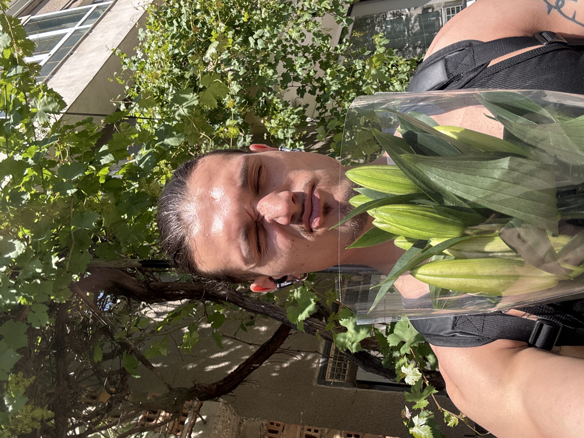 A close-up, selfie-style shot features a young man with dark hair pulled back, squinting his eyes and puckering his lips as he holds a bouquet of green lily buds wrapped in clear plastic. He is wearing black earbuds and a silver hoop earring in his left ear. A black backpack strap is visible over his left shoulder. The background is dominated by lush green foliage, with hints of a building and a vine-covered wall suggesting an outdoor urban setting, possibly in Sofia, Bulgaria, given the architectural style and dense greenery. Sunlight casts shadows, indicating a bright day.