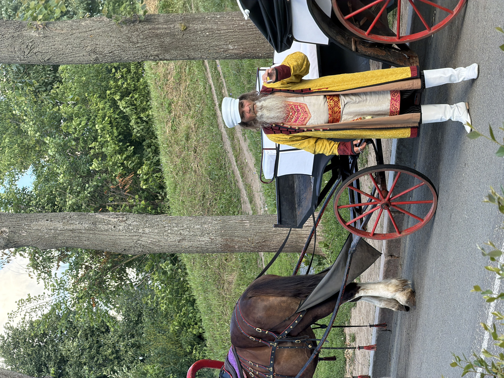 A man in traditional clothing stands next to a horse-drawn carriage in Suzdal, Russia. The man is wearing a white cap, a yellow robe with red trim, a white tunic with red embroidery, a red sash, striped pants, and white boots. He has a long beard and is holding a small object in his right hand. The horse is brown and is harnessed to the carriage, which has white seats and red wheels. The scene is set outdoors with trees and a grassy hill in the background. The ground is paved with asphalt.