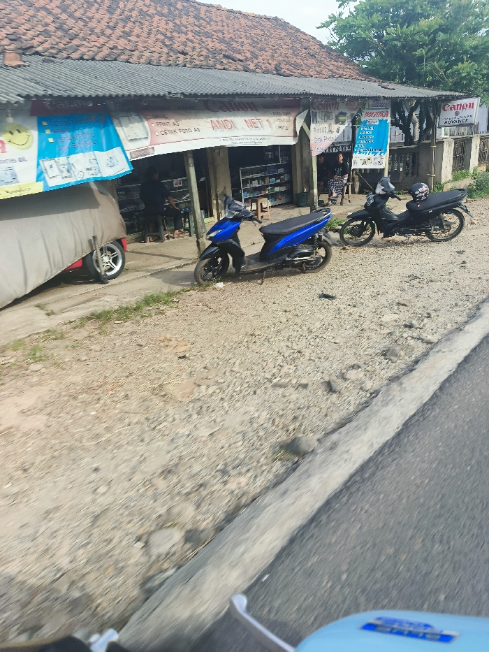The image shows a street in Sukatani, Indonesia (latitude: -6.608, longitude: 107.414). There are two parked motorcycles, one blue and one black, in front of a shop with a sign that reads "Andi Net 1". The blue motorcycle is parked closer to the shop while the black one is parked further away. There are two people standing in front of the shop and a man sitting on a chair next to a window. The shop has a sign that reads "Canon". The shop is made of brick and has a tiled roof. There is a gray tarp covering a portion of the shop. There is a patch of grass next to the shop and a paved road in front of the shop and motorcycles.