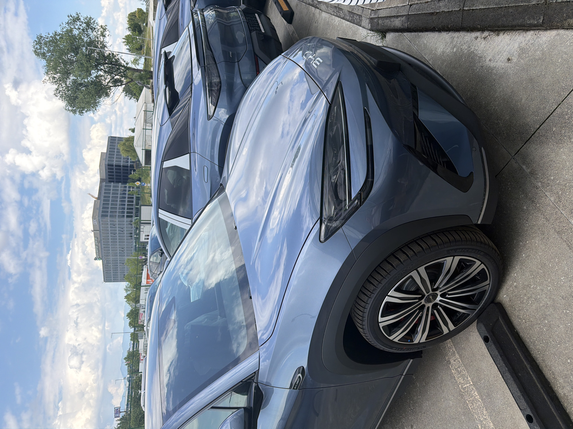An outdoor, daytime shot shows a dark blue-grey modern SUV parked on a concrete surface. The car, in the foreground, features distinctive multi-spoke wheels with a "BYD" logo on the hub cap and "MICHELIN" visible on the tire. The text "CH3" is faintly visible on its hood. Clouds are reflected prominently on the car's glossy bodywork. Behind it, several similar vehicles are parked in a row.
In the background, a large green tree stands against a bright blue sky dotted with white clouds. Further back, a multi-story modern building is visible, with "IBM" and "SOFIA AIRPORT CENTER" clearly readable on its facade, indicating a commercial area. Distant mountains or hills can be seen on the horizon. The setting appears to be a parking lot or a car display area in Busmantsi, Bulgaria, under sunny weather conditions. No people or specific activities are depicted.