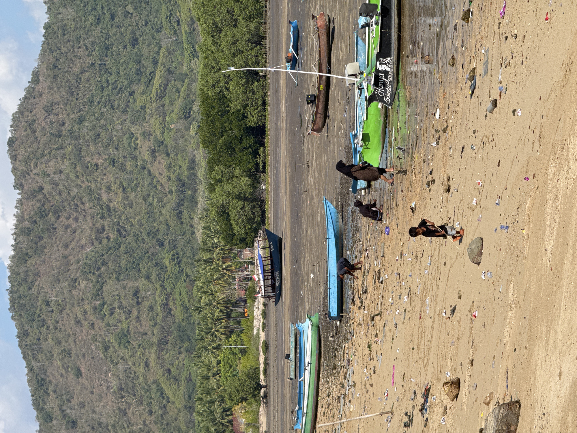 In Kertasari, Indonesia, a wide, sandy beach is littered with debris. Several boats are grounded on the muddy shoreline, some partially submerged in shallow water. In the foreground, a young boy in a black t-shirt and orange flip-flops is playing with a stick. Further back, a woman wearing a headscarf and a long brown dress is walking, while another person kneels near a green boat. In the background, a dense, green forest meets a dry, mountainous landscape under a partly cloudy sky.
