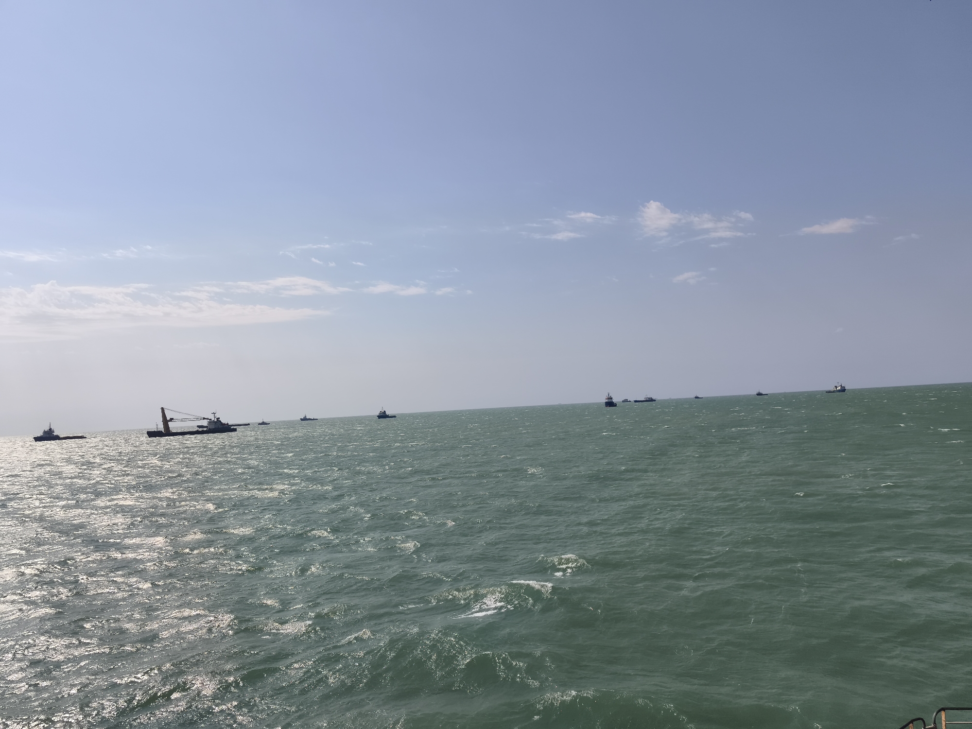 The image depicts a wide expanse of the Caspian Sea under a clear, light blue sky with sparse white clouds. The water is a deep turquoise green, with visible waves and sunlight glinting off its surface. Several vessels are scattered across the horizon. On the left side of the image, a large barge with a crane is prominent, accompanied by smaller tugboats. Further out, other ships, likely cargo vessels, are visible as silhouettes against the horizon. The scene appears to be a calm day at sea, with no people visible and no specific actions or interactions taking place beyond the presence of the ships. The location is consistent with the coastline of Pirallahi Raion, Azerbaijan, which is known for its industrial port activities and offshore platforms.