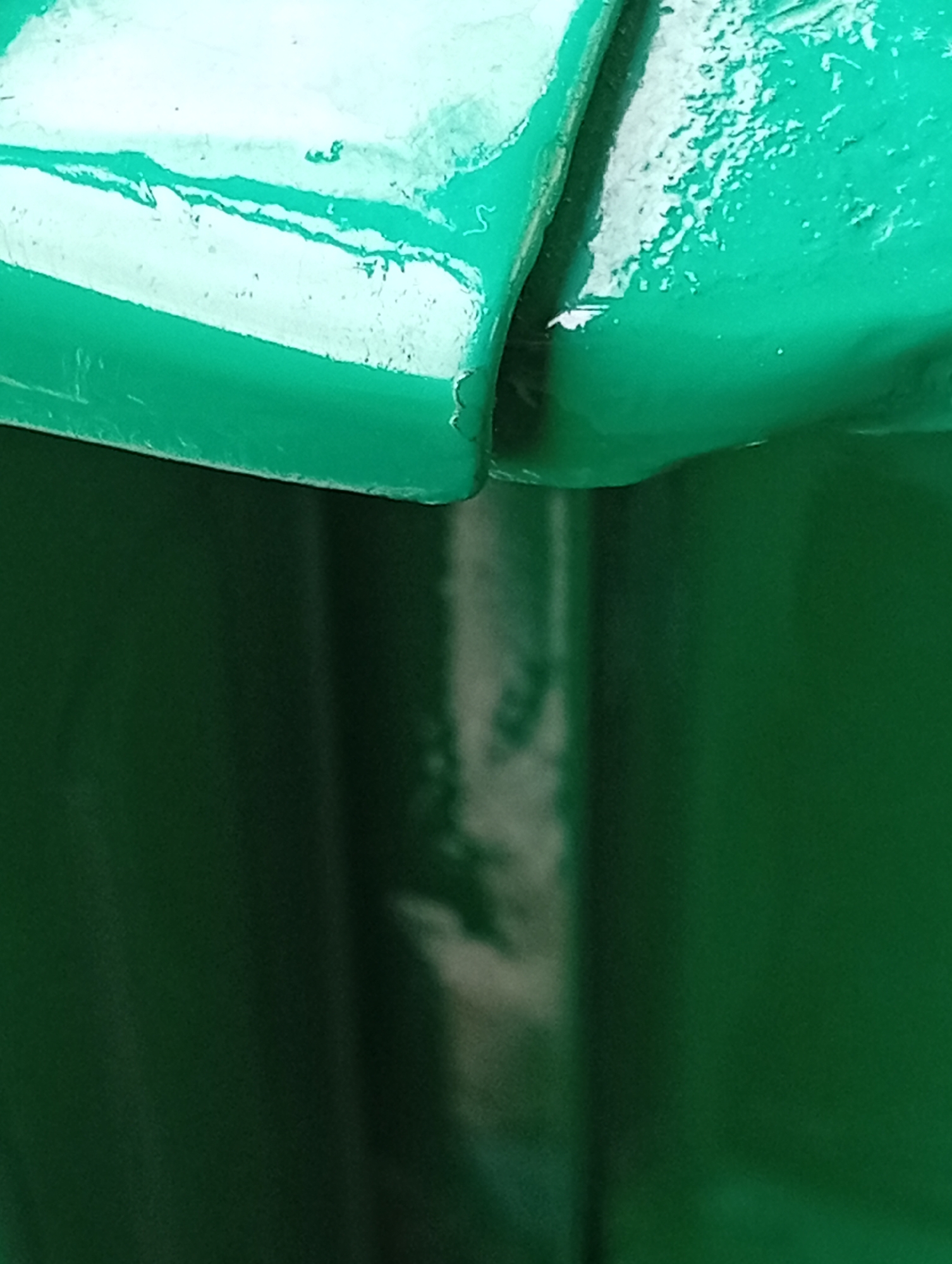 The image shows a close-up, abstract view of a metallic structure, likely painted green. The surface exhibits a glossy finish with some textured imperfections, possibly due to weathering or manufacturing. The lighting suggests an outdoor environment, with highlights and shadows indicating the form of the object. The texture appears uneven in places, with subtle scuff marks or debris on the surface. The deep green color dominates the frame, with a lighter, almost white area in the upper portion of the image. The perspective is tight, focusing on the details of the material and its surface. There are no people, animals, or discernible urban landscape elements visible in the image.