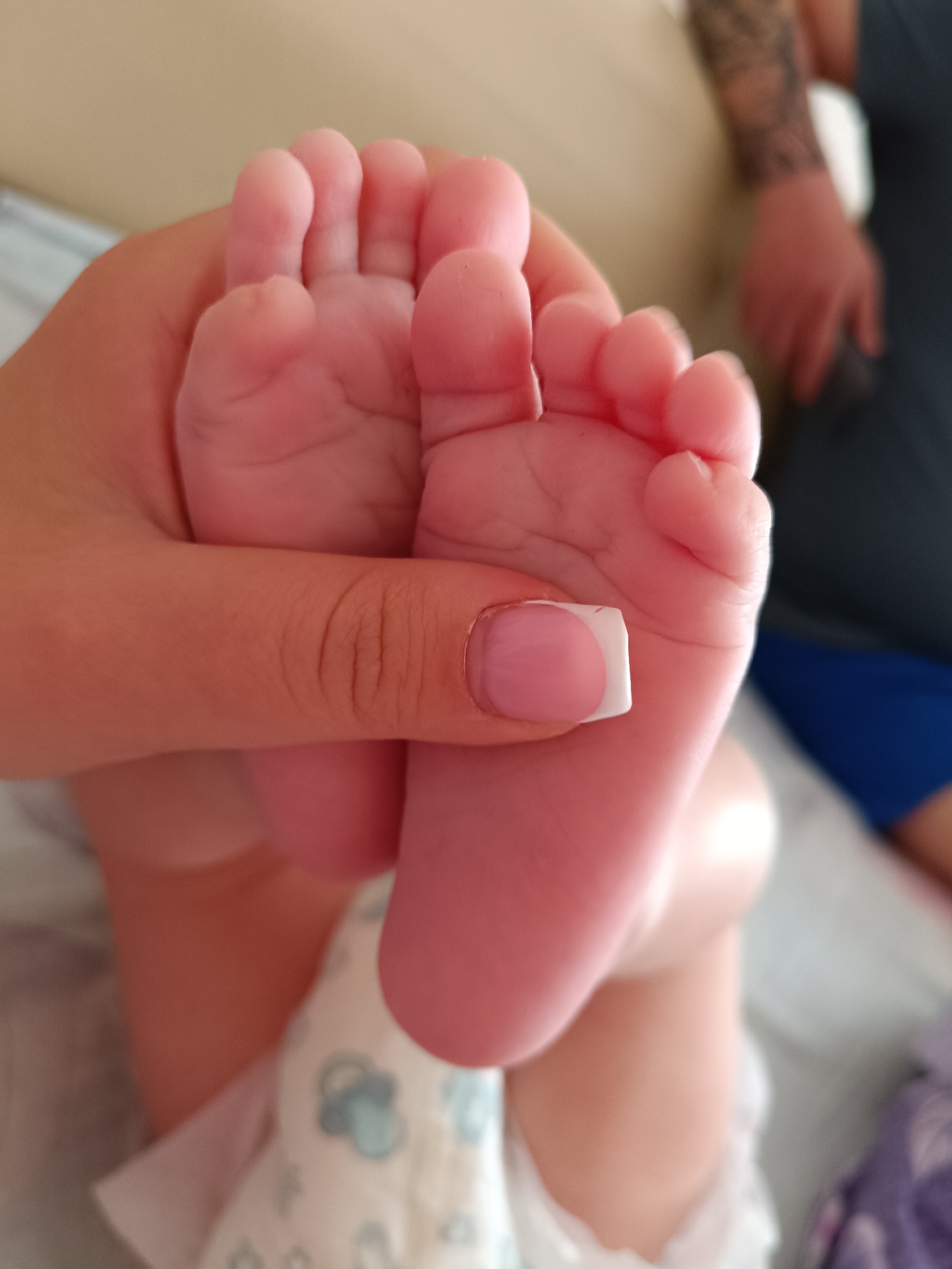 A person with manicured fingernails is holding a newborn baby's feet. The baby is wearing a diaper with a light blue and grey cloud pattern. In the background, a person with a tattooed arm is visible. The location is unknown and cannot be determined from the image.