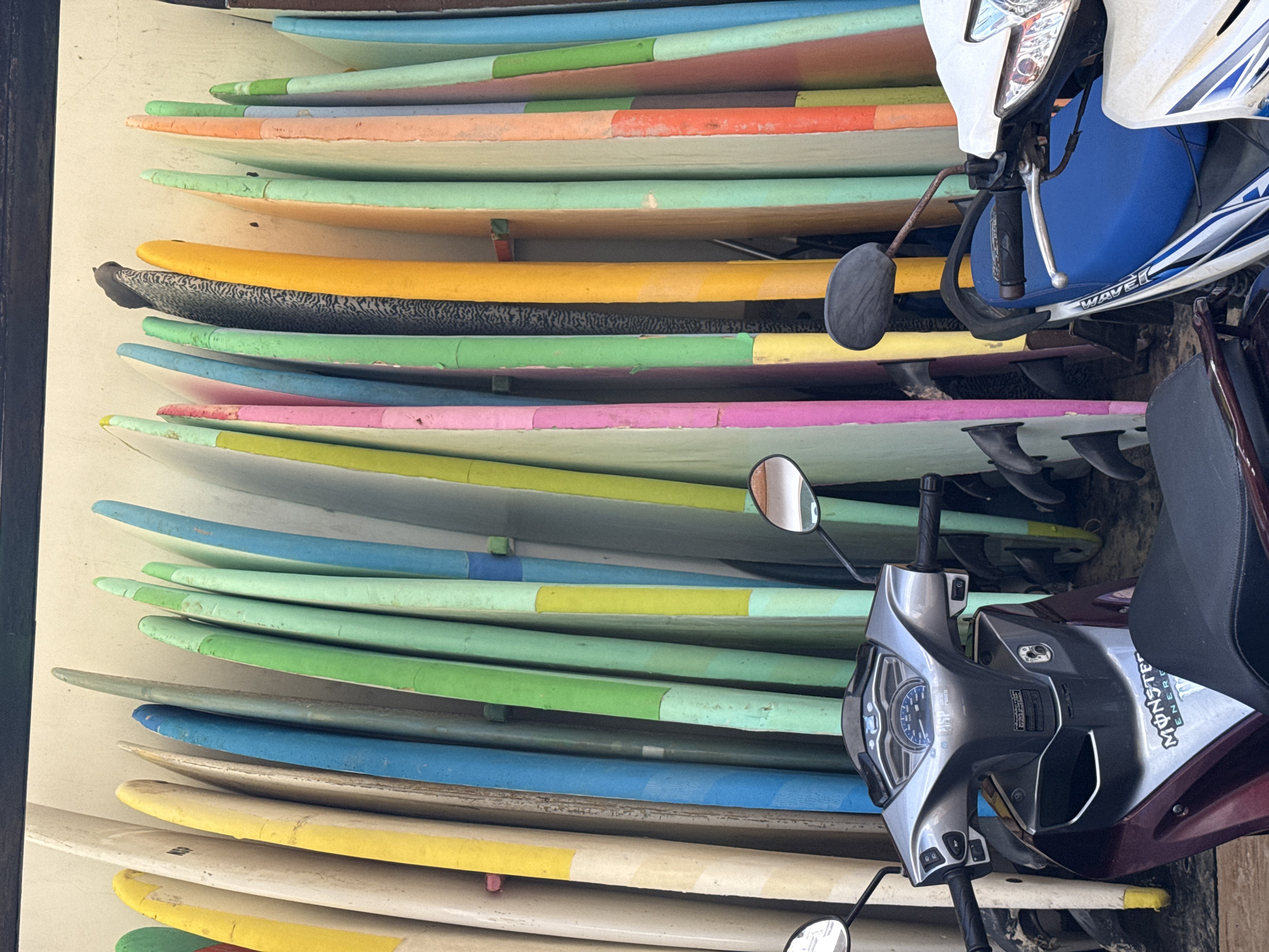 In Karon, Thailand, a rack of colorful surfboards stands against a wall. The surfboards are of various colors, including yellow, white, blue, green, pink, and orange. Some of them have black fins visible at their bases. In the foreground, two motorcycles are parked. The motorcycle on the left is dark red with a silver front panel and black seat. The motorcycle on the right is white with a blue seat and silver handlebars. Both motorcycles appear to be stationary. The environment suggests an outdoor setting, possibly near a beach.