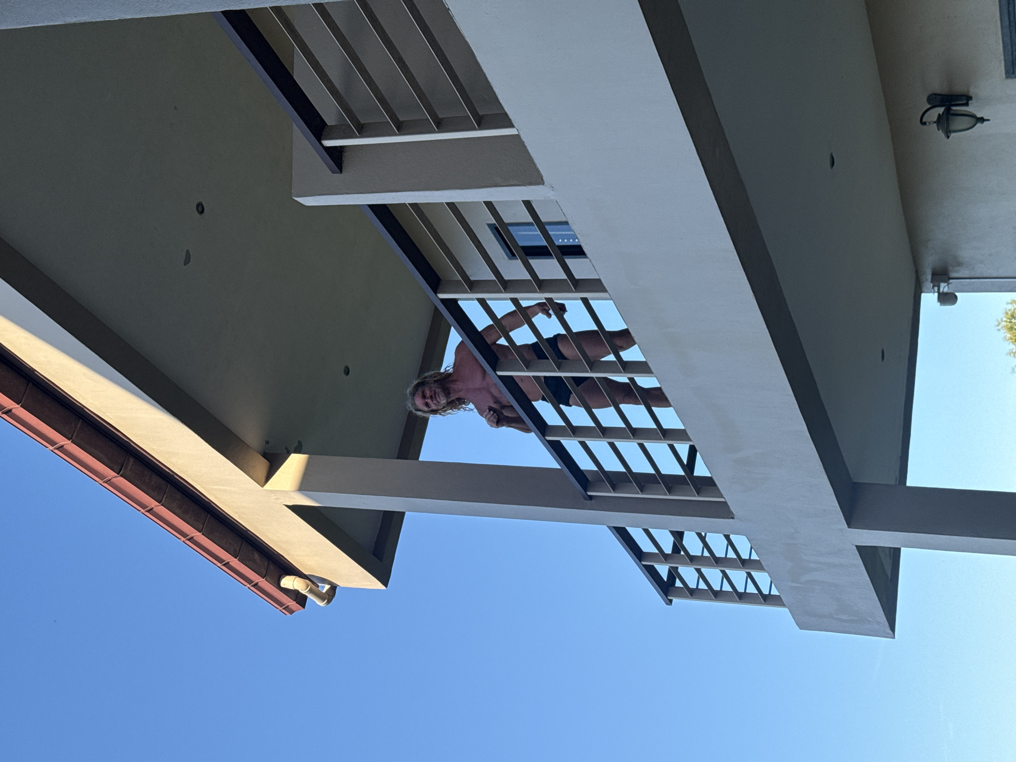 In Baan Chaweng, Thailand, a man with long blond hair stands on a balcony, looking to the right. He is shirtless and wearing black swimming trunks. The balcony has a white railing with dark horizontal bars. The building's architecture features a tiled roof with a reddish-brown hue and a light-colored exterior. The sky above is a clear blue. The perspective is from below, looking up at the man and the building.